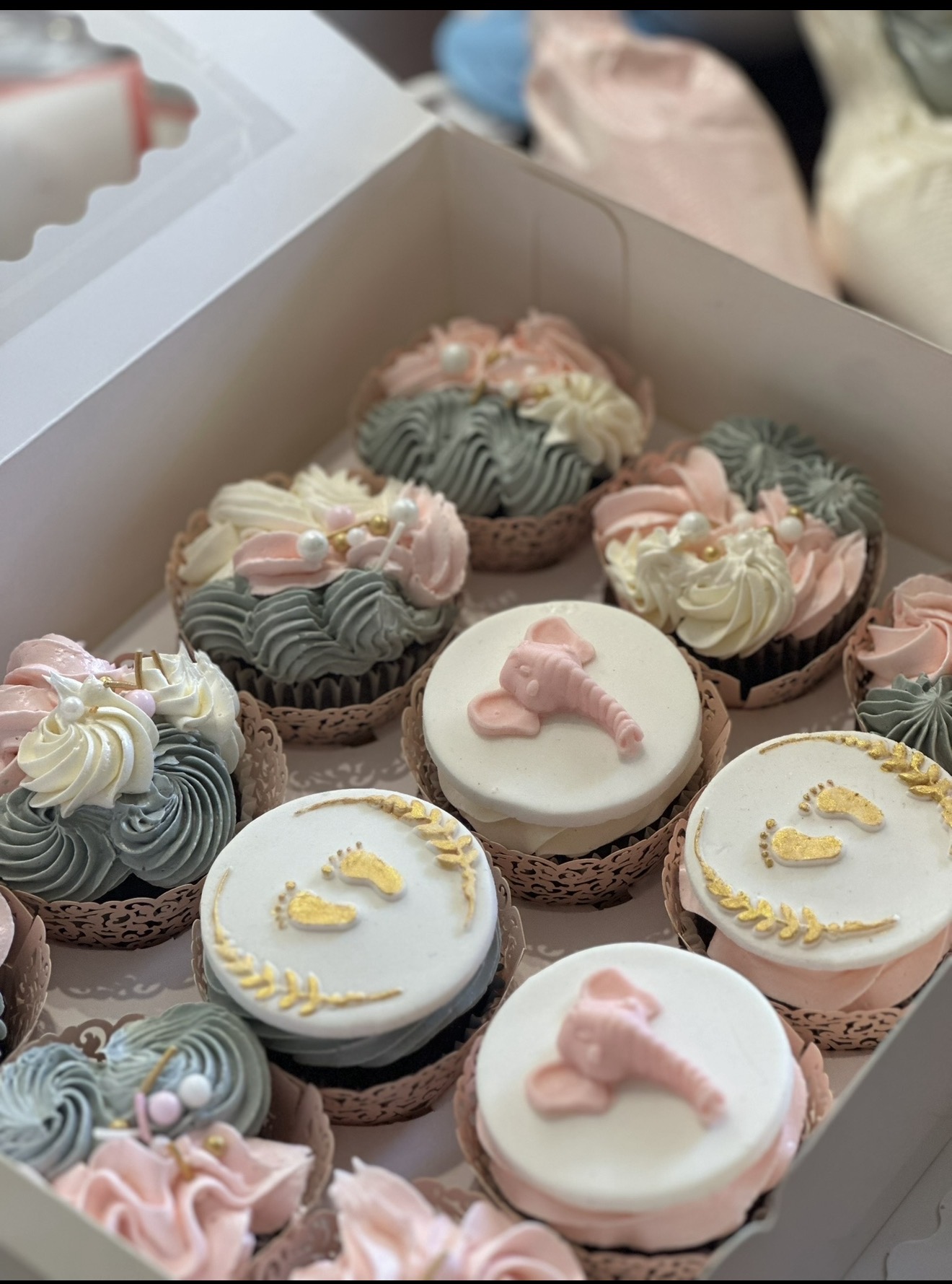 Box of decorated cupcakes with pink, white, and gray icing, some with animal designs, arranged in a white box.