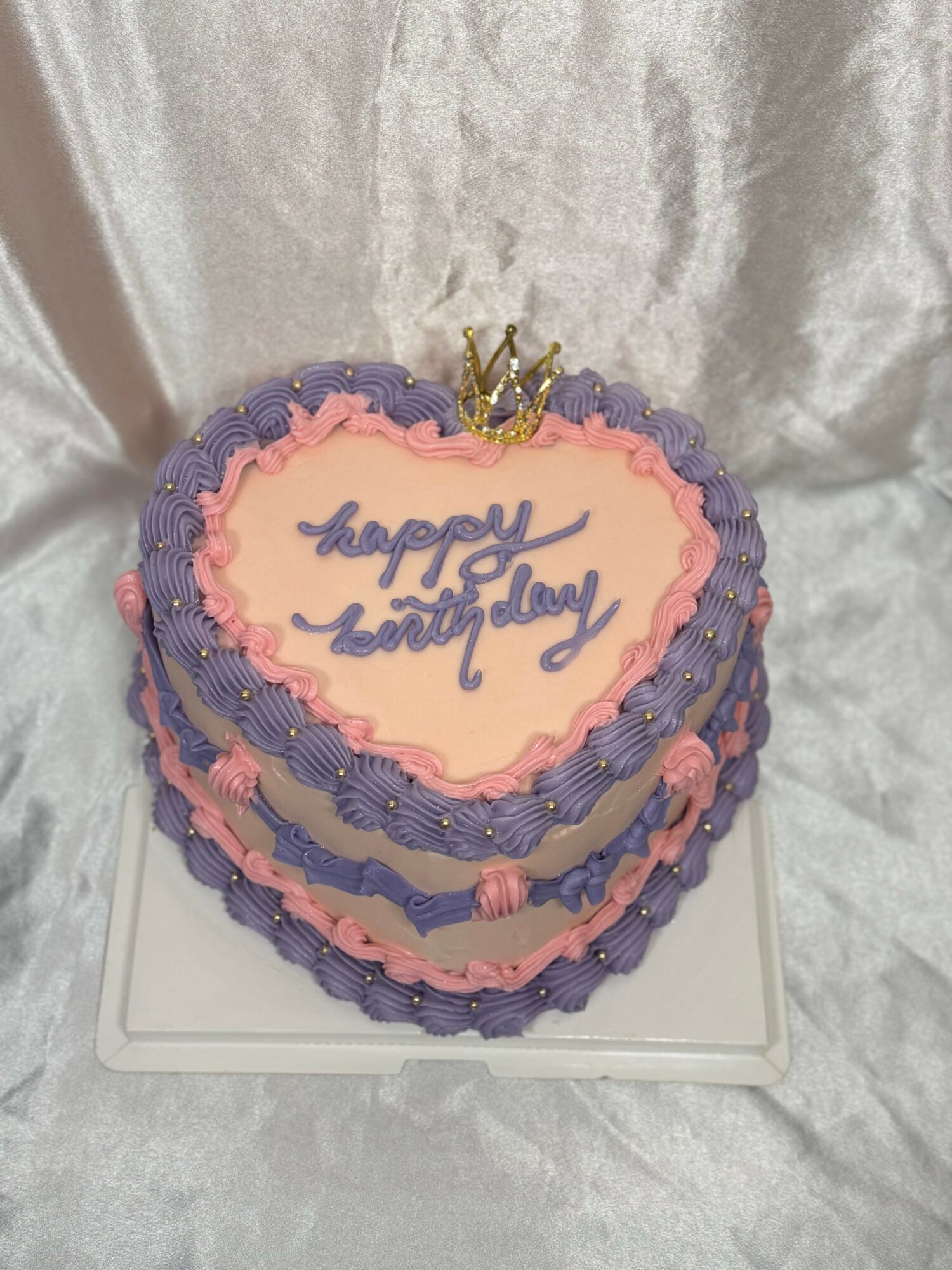 Heart-shaped birthday cake with purple and pink icing, a small gold crown on top, and the message 'happy birthday' written in blue.