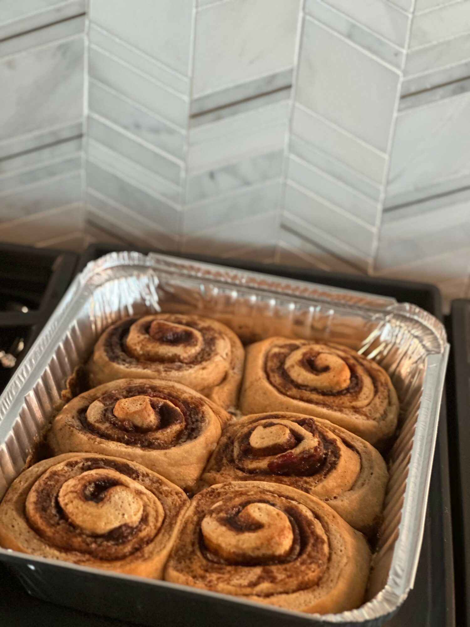 Six cinnamon rolls in a foil tray on a kitchen counter with white tiled backsplash.