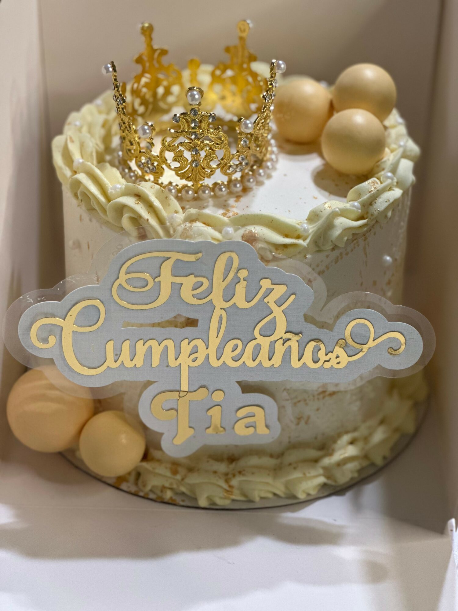 Birthday cake with a gold crown, cream decorations, and a sign reading 'Feliz Cumpleaños Tia'.