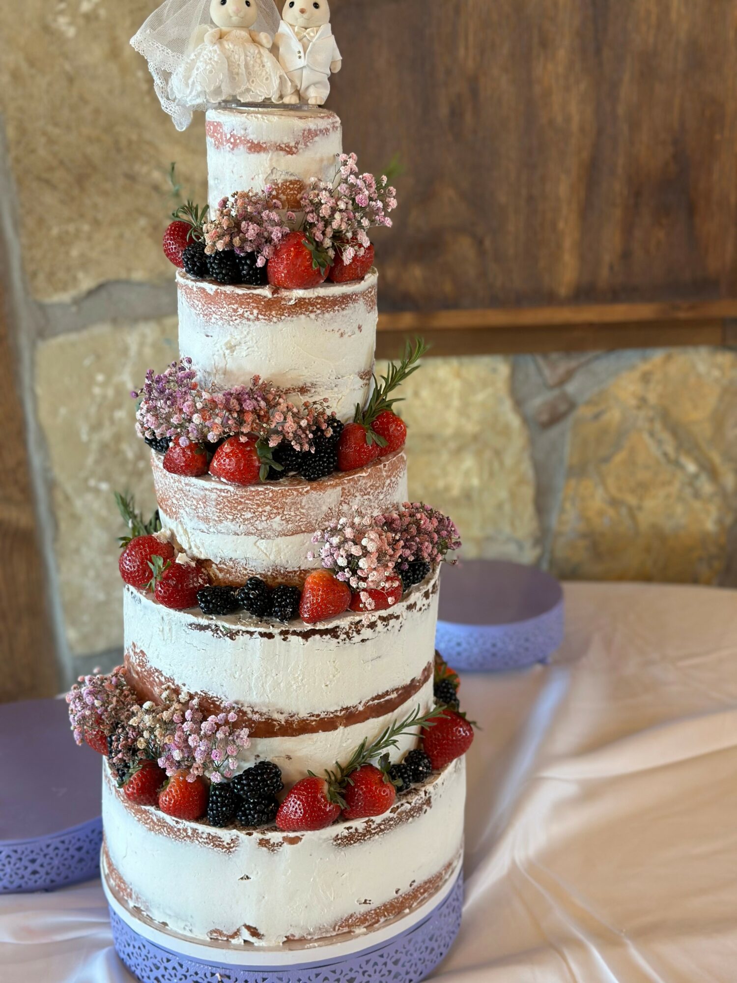 Tall, multi-layered cake decorated with berries, flowers, and small figurines on top, set against a stone and wood background.