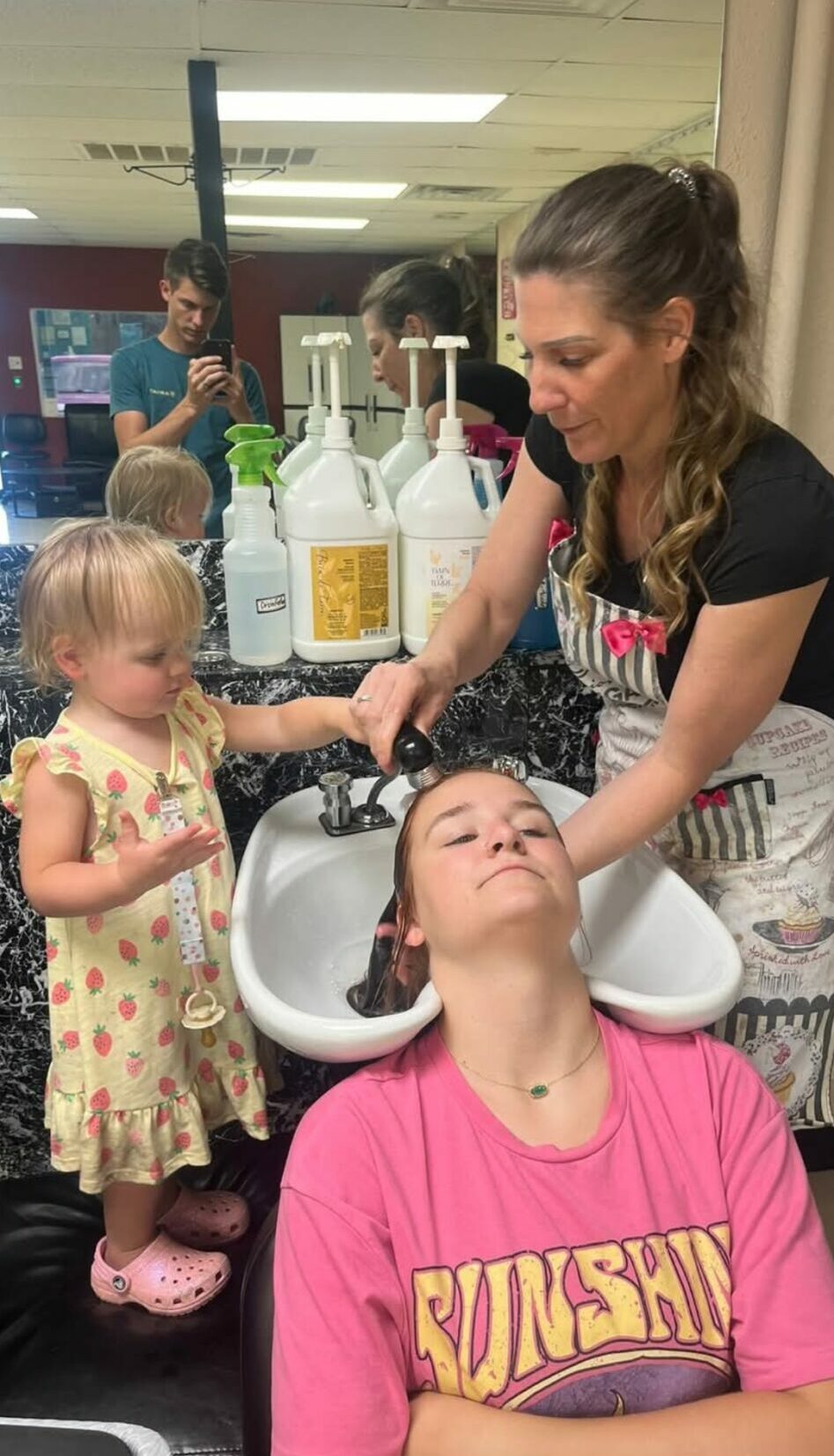 Woman washing a child's hair in salon sink, young girl with pink shirt, another child in floral dress watching, salon background.