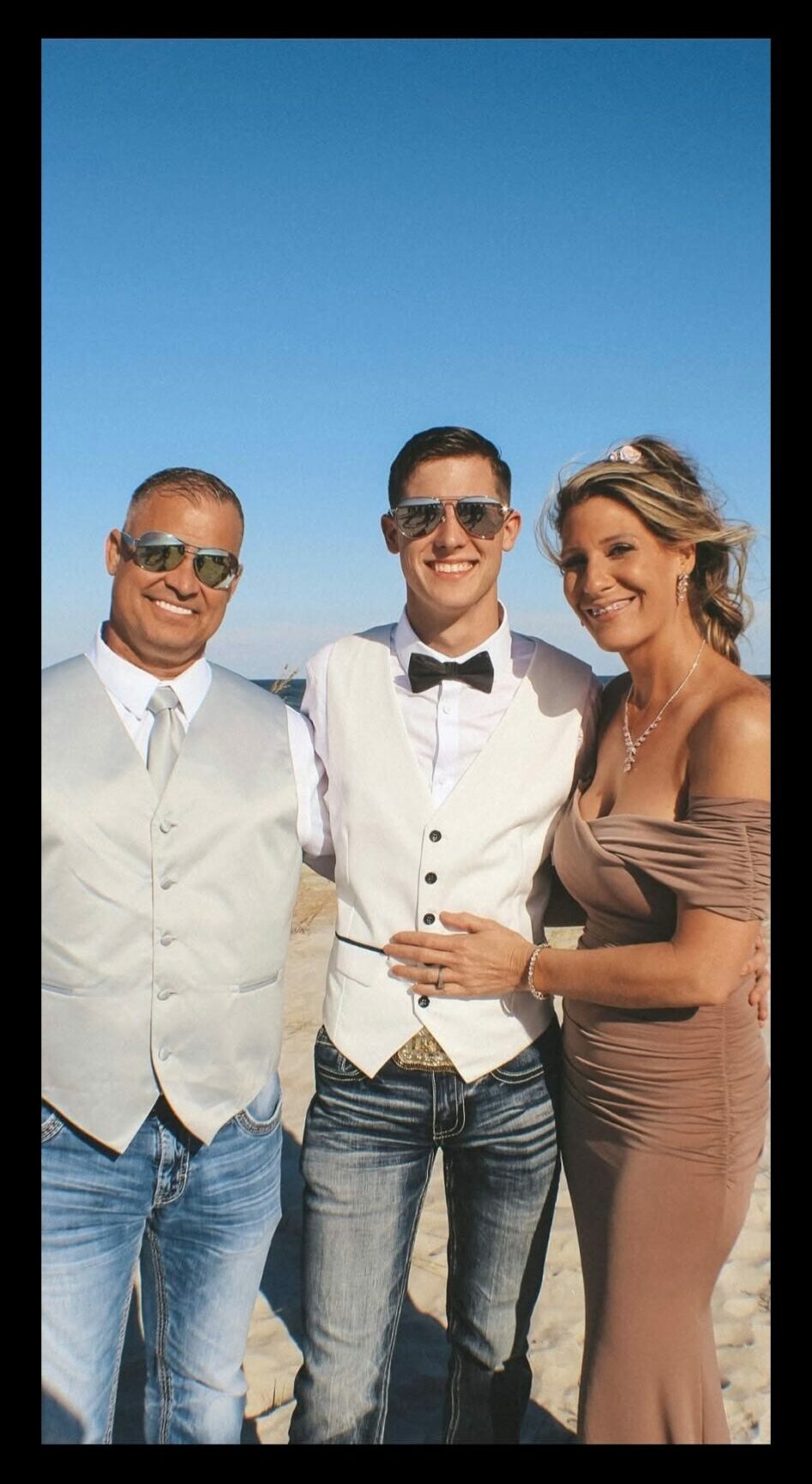 Three people smiling outdoors against a clear blue sky, dressed in formal and casual attire.