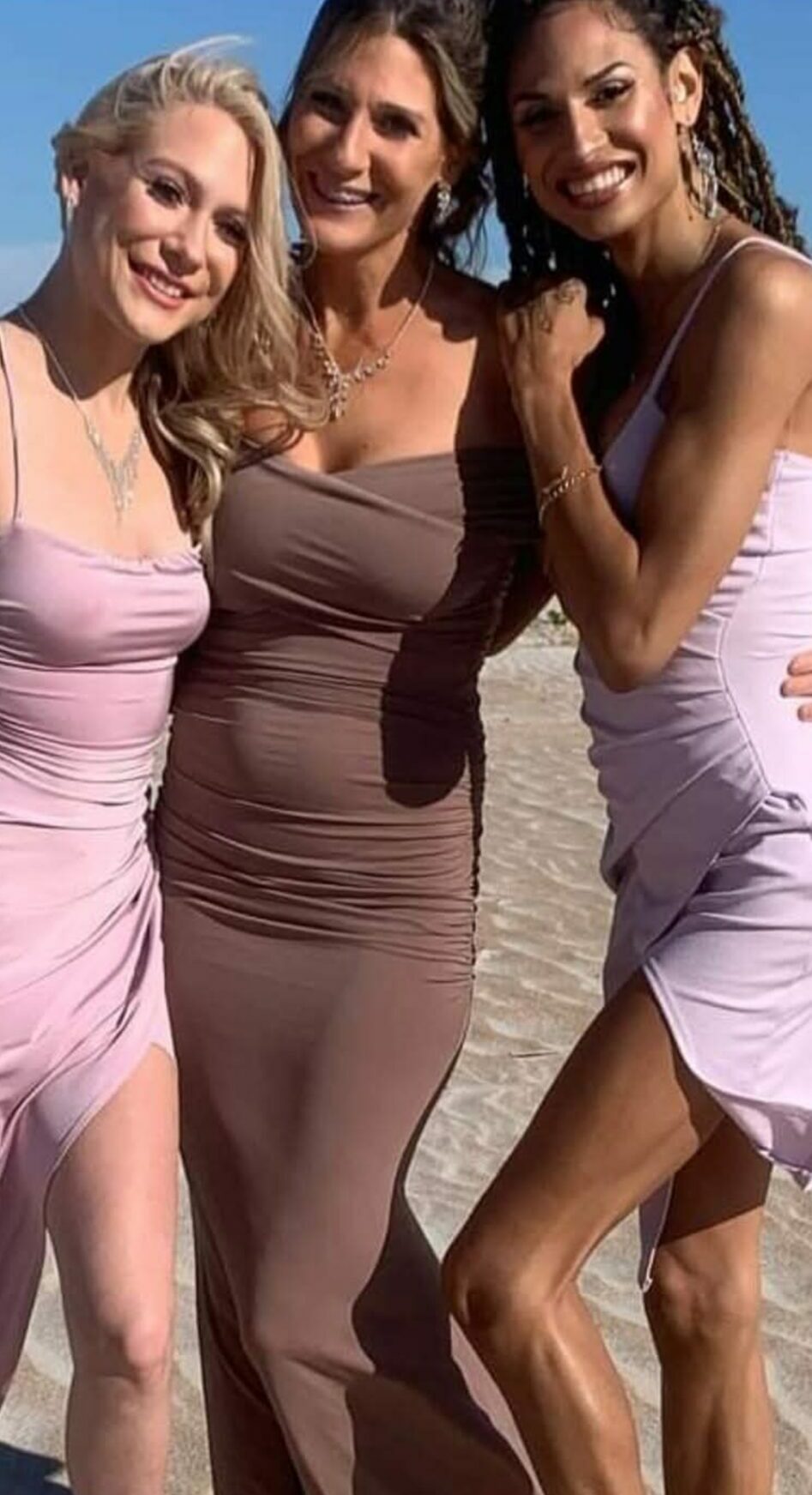 Three women standing close together outdoors, smiling, with a beach and sky in the background.