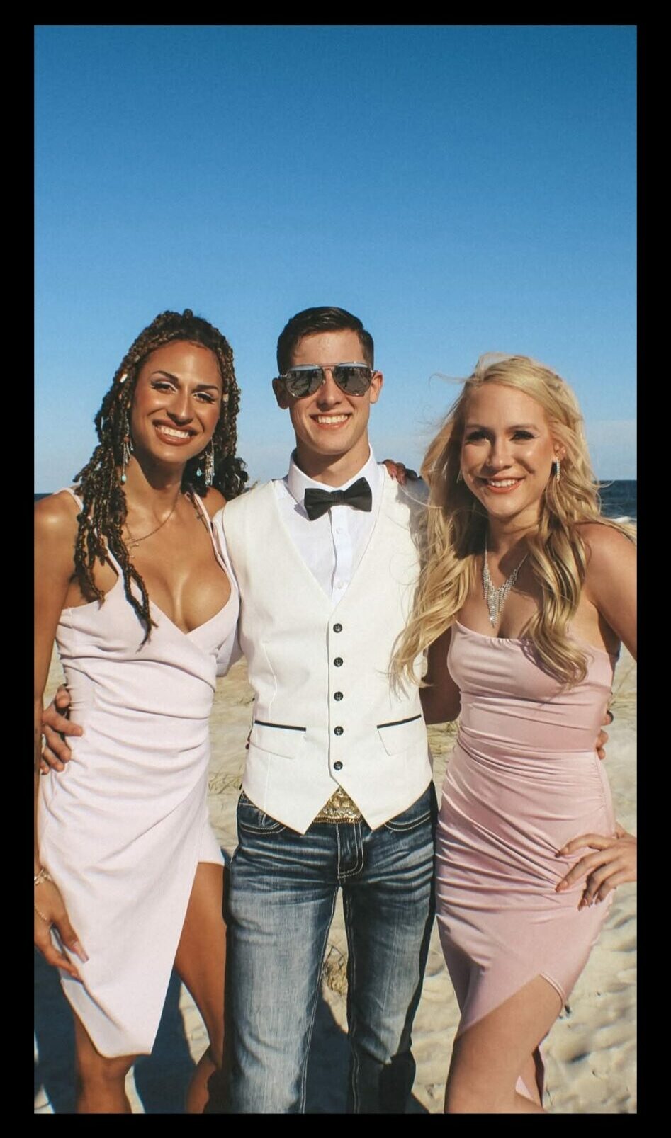 Three people standing outdoors against a clear blue sky, smiling, with two women in pink dresses and a man in a white shirt and bow tie.