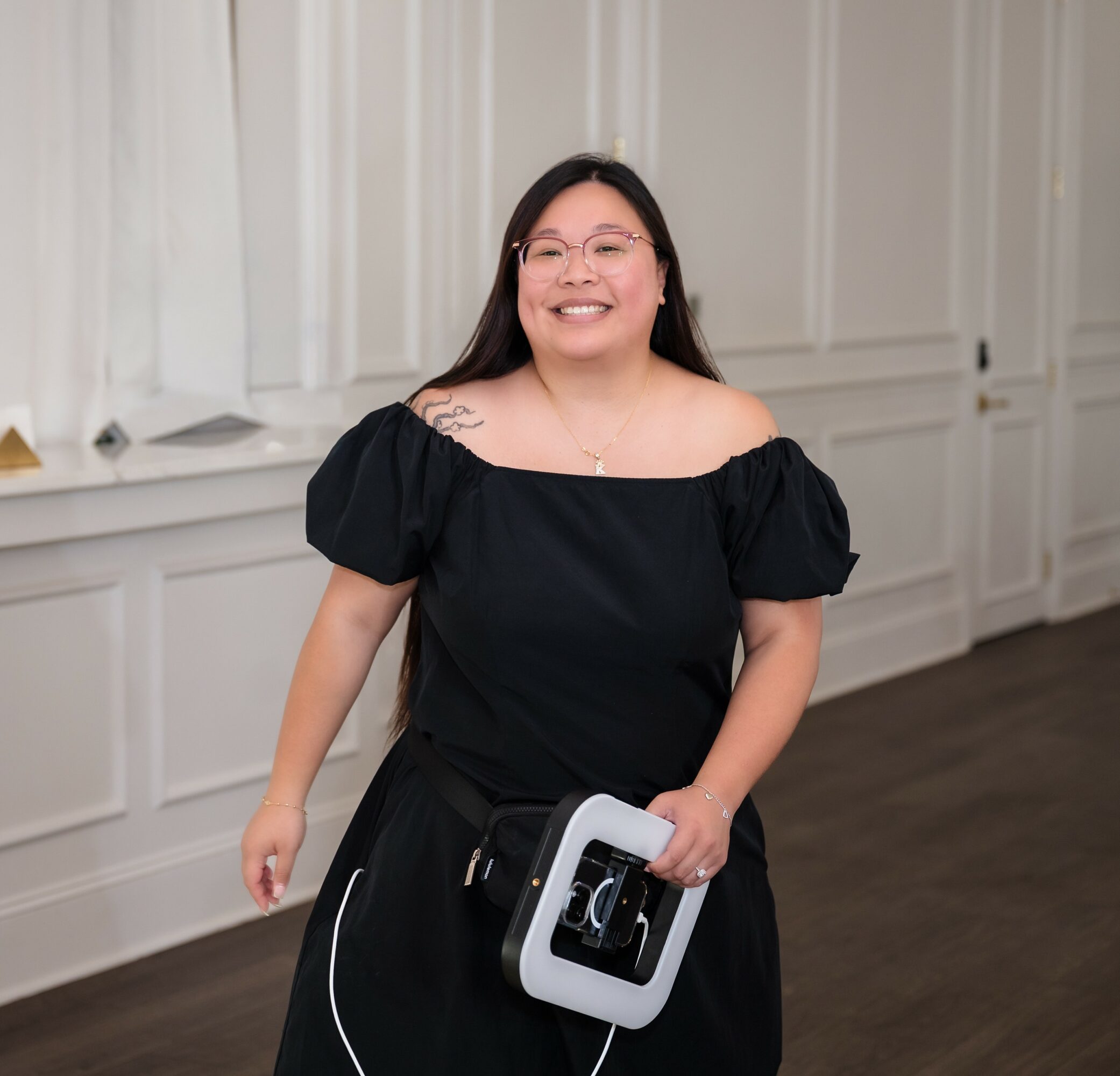 Smiling woman with glasses in a black off-shoulder dress holding a device, standing in a room with white panel walls.