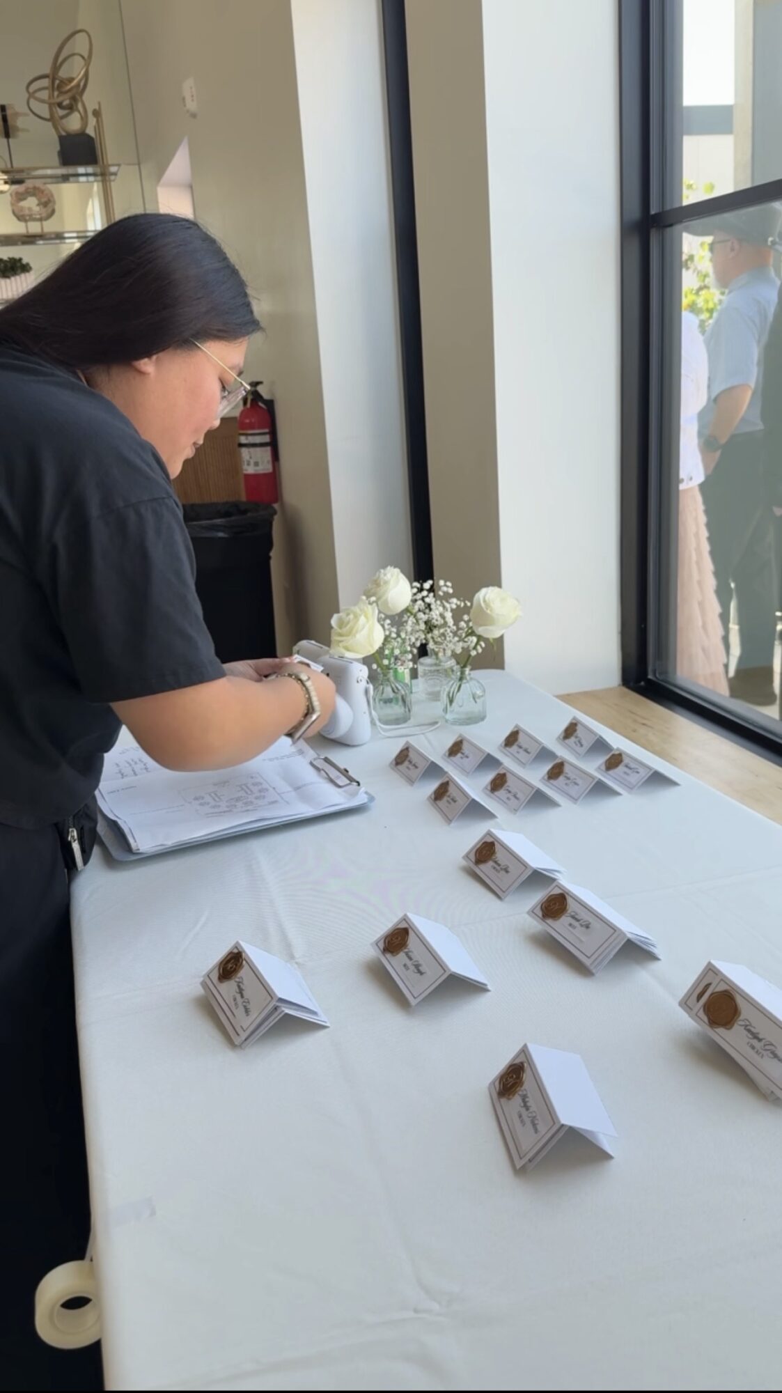 Person arranging small display cards on a table near a large window, with flowers and a fire extinguisher nearby.
