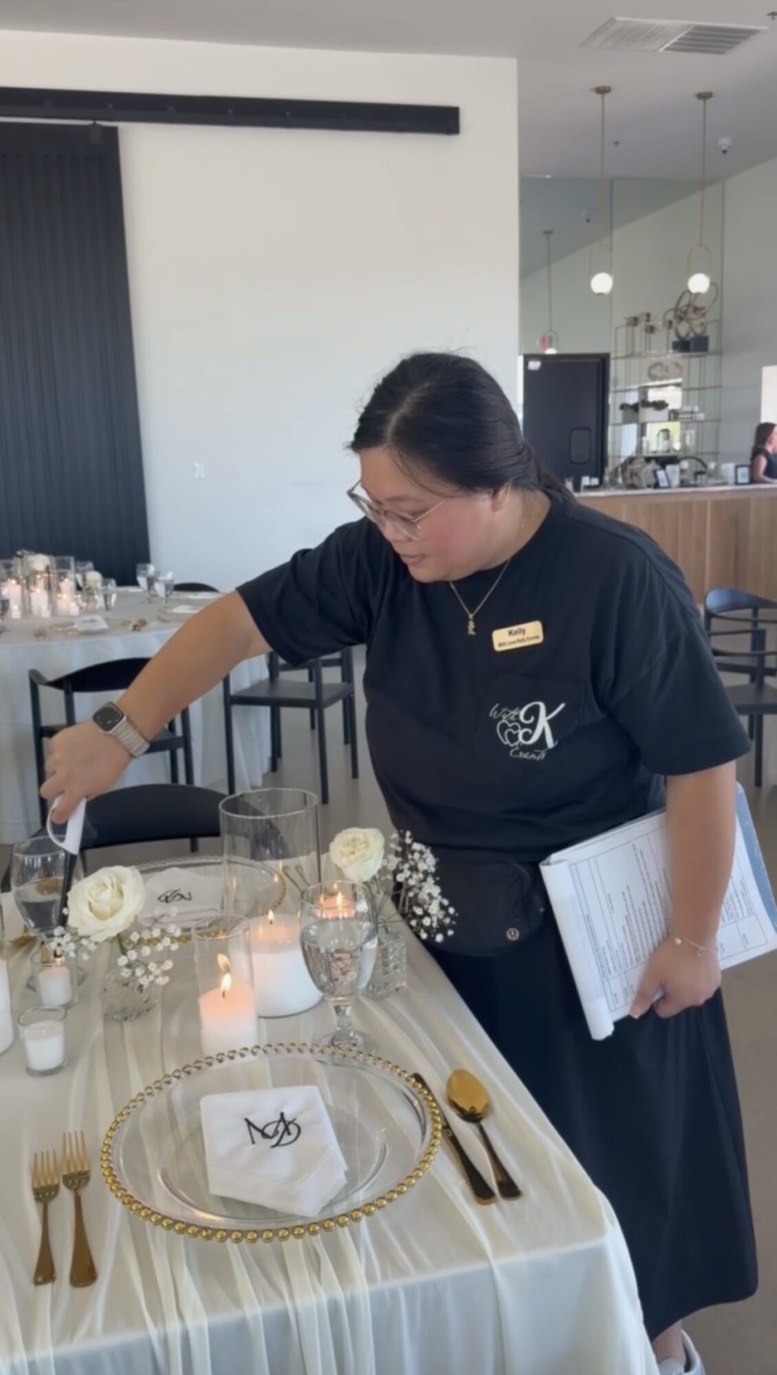 Woman setting a table with white roses, candles, and gold utensils in a modern restaurant interior.