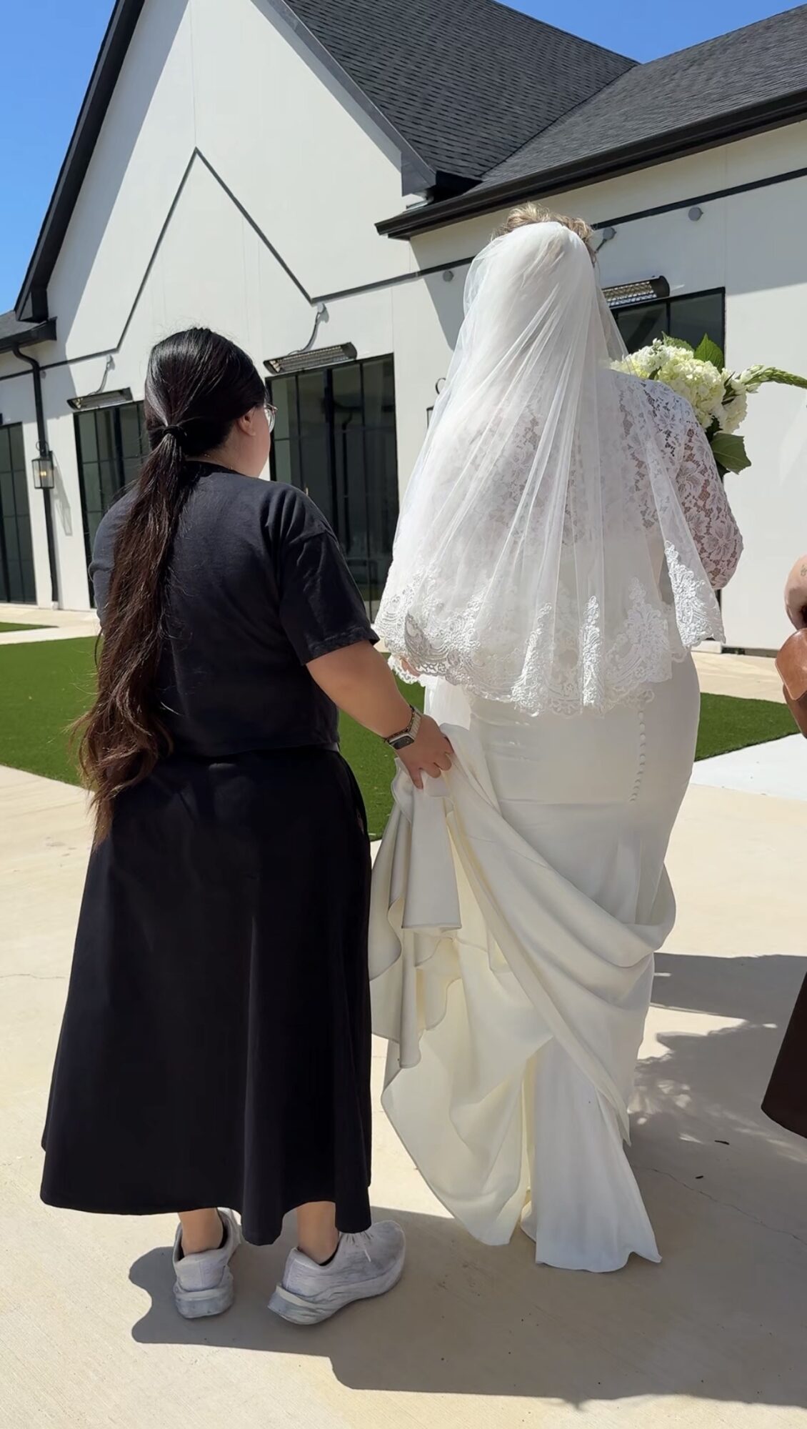Two women, one in a black dress and the other in a white wedding gown with a veil, walking outside near a building.