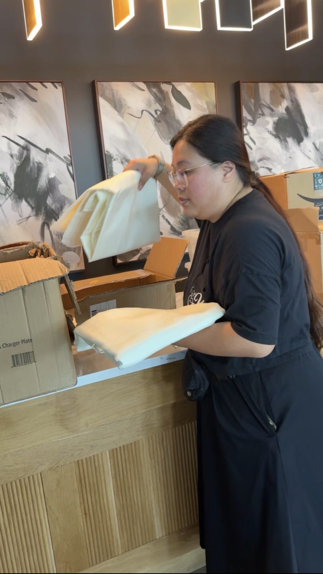 Woman with glasses and dark hair at counter, handling paper or packaging materials, in a modern store or cafe setting.