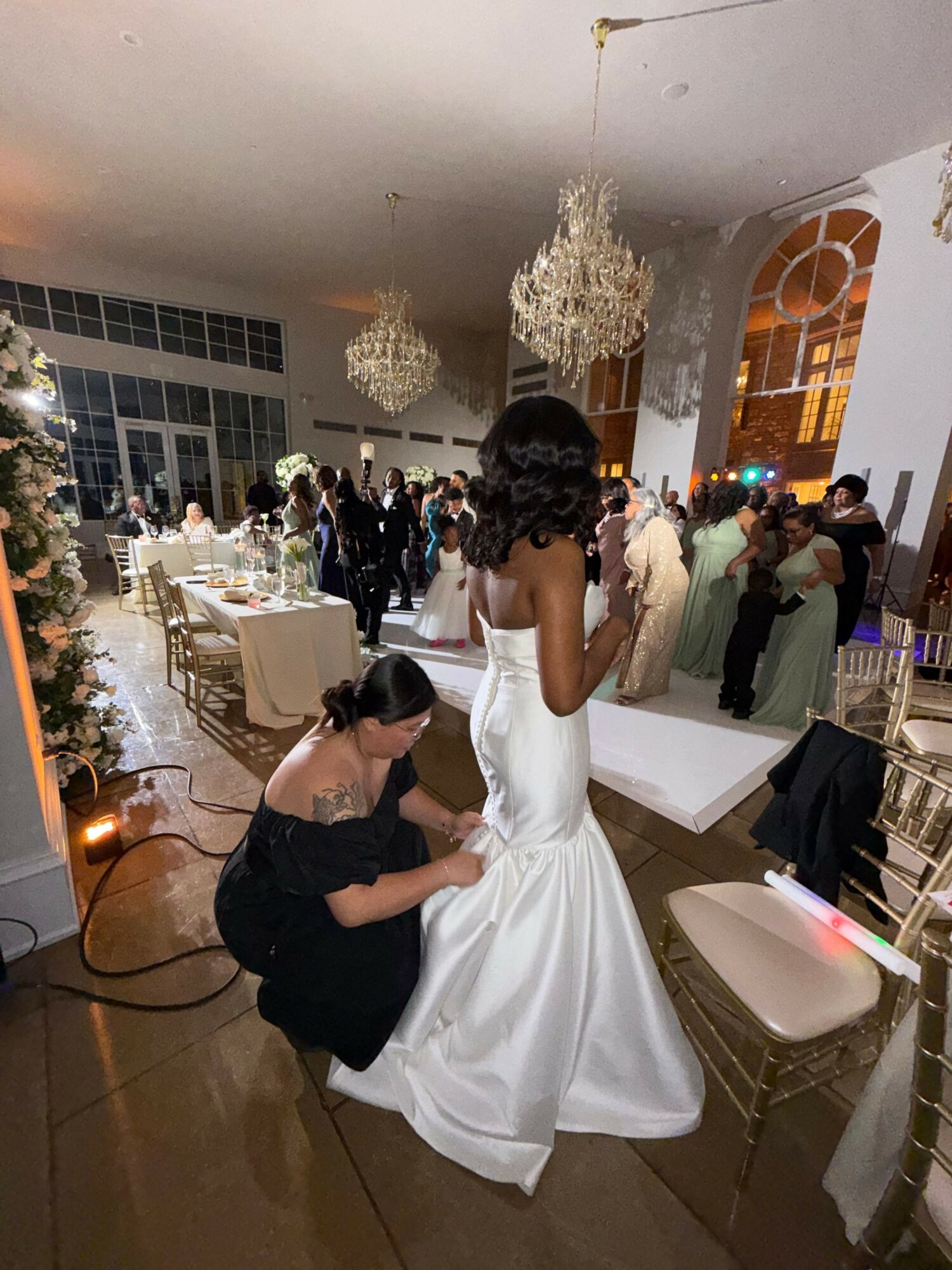 A woman in a white dress and a woman in black are seated at a table during a celebration, with guests and decorations in the background.