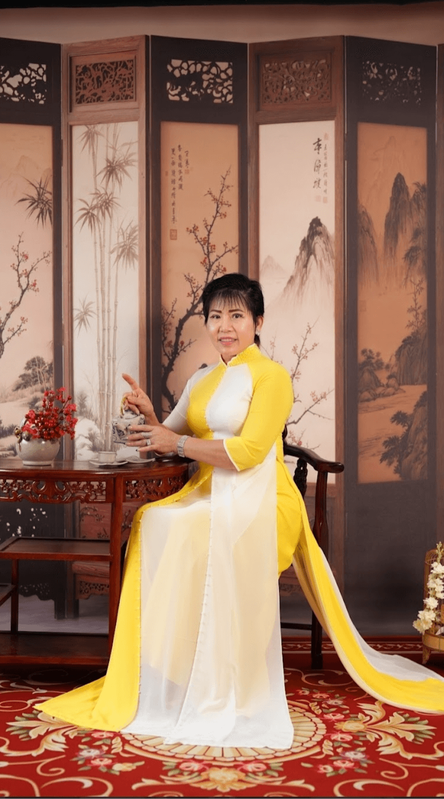 Woman in traditional dress sitting on a wooden chair with a folding screen behind her, holding a small object, on a red patterned carpet.