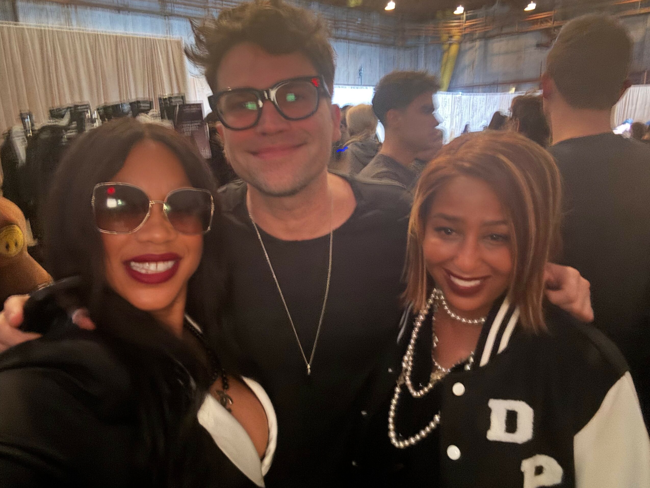 Three people smiling at a crowded indoor event, two women and one man, with the women wearing sunglasses and the man wearing glasses.