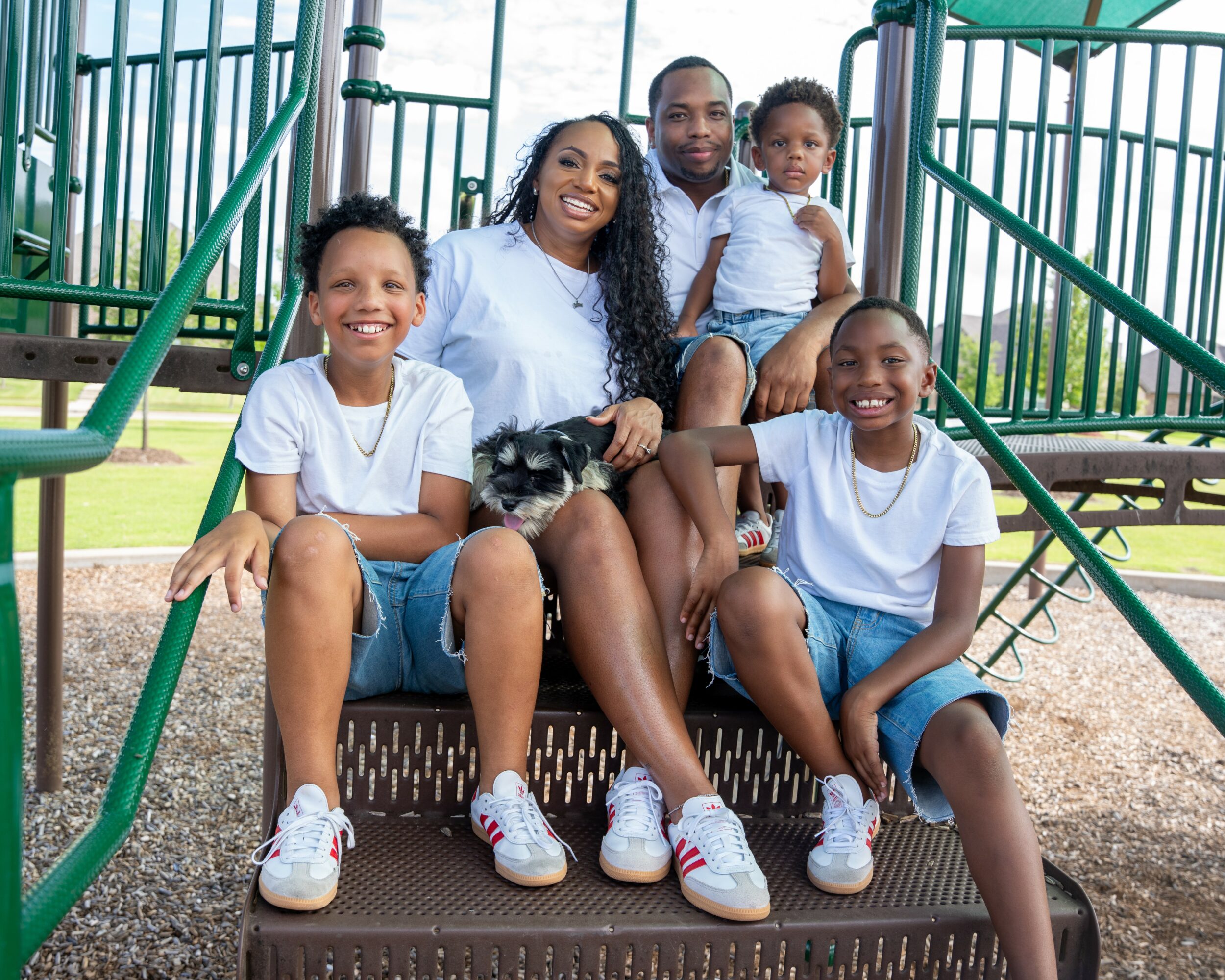 Group of five people, including children and an adult, sitting on playground equipment outdoors, smiling, with a dog.