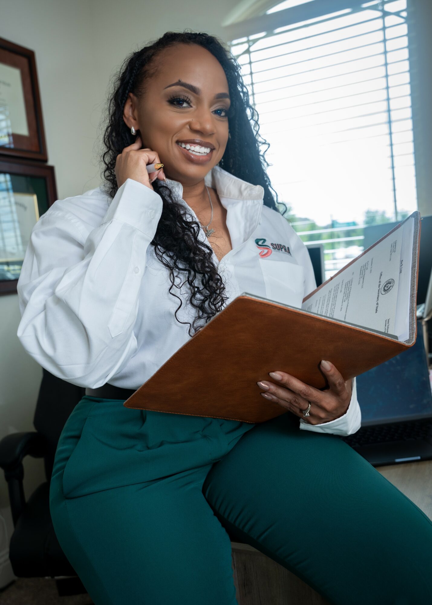 Woman smiling, holding a folder, sitting in an office with window blinds behind her.