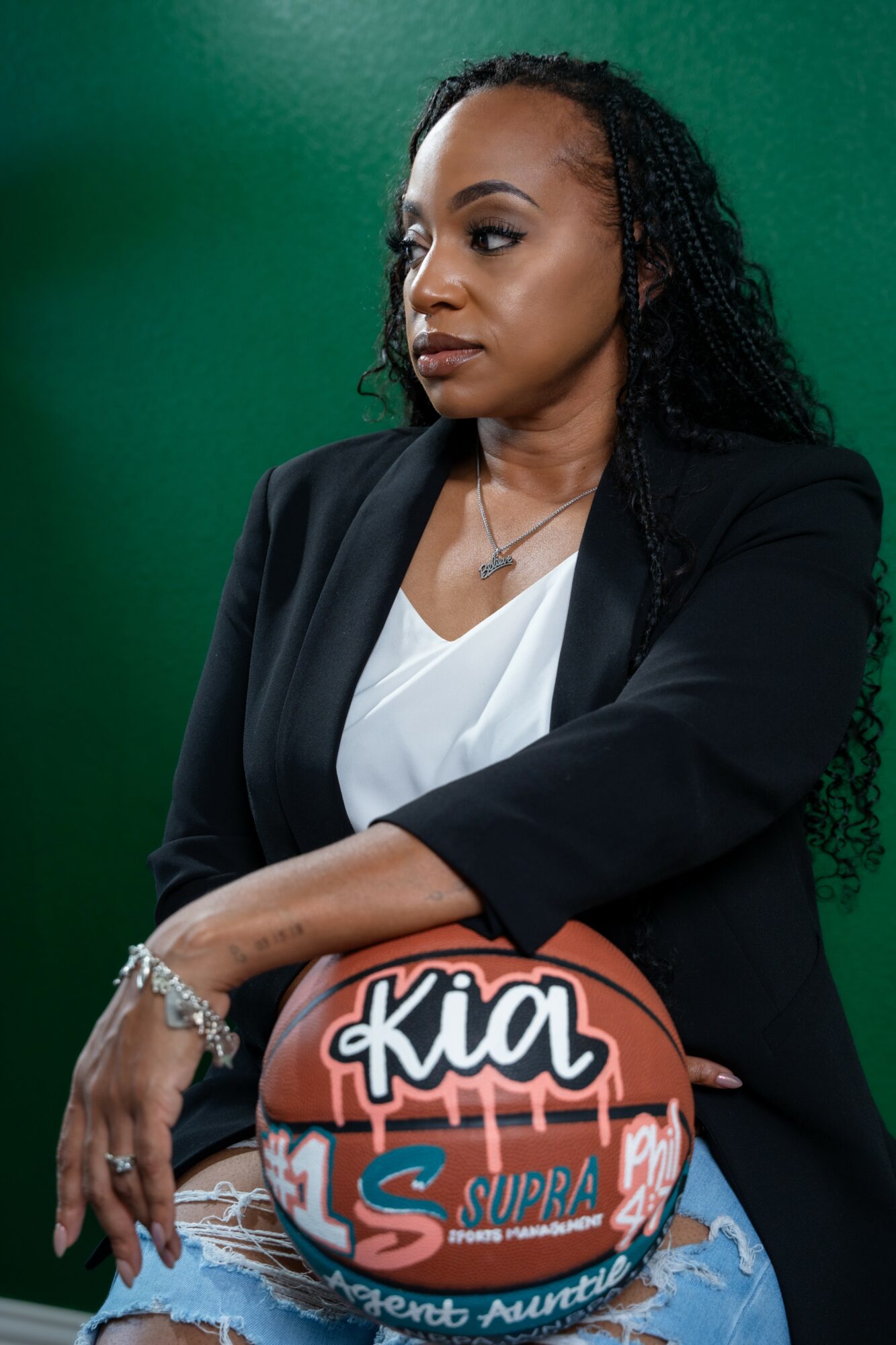 Woman with long curly hair in a black blazer and white top holding a basketball against a green background.