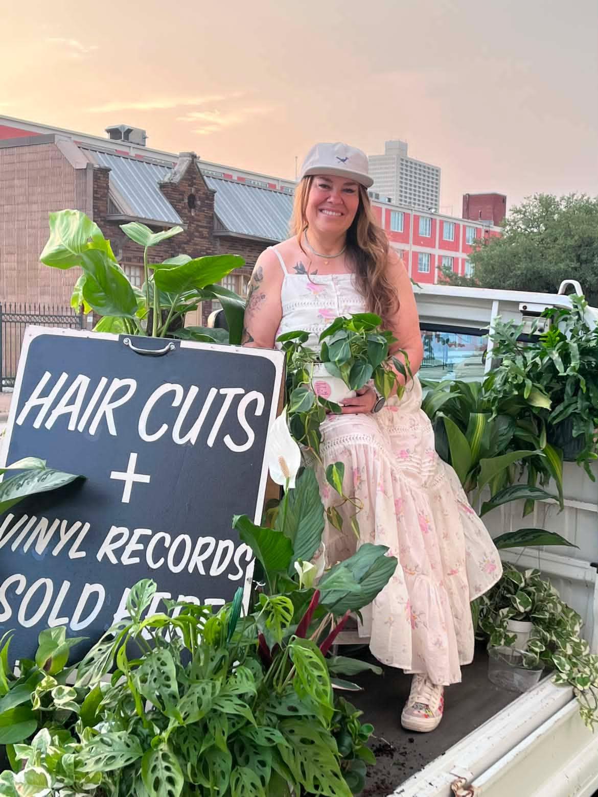 Woman with long hair wearing a hat and dress, sitting among plants on a rooftop, with a sign about hair cuts and vinyl records.