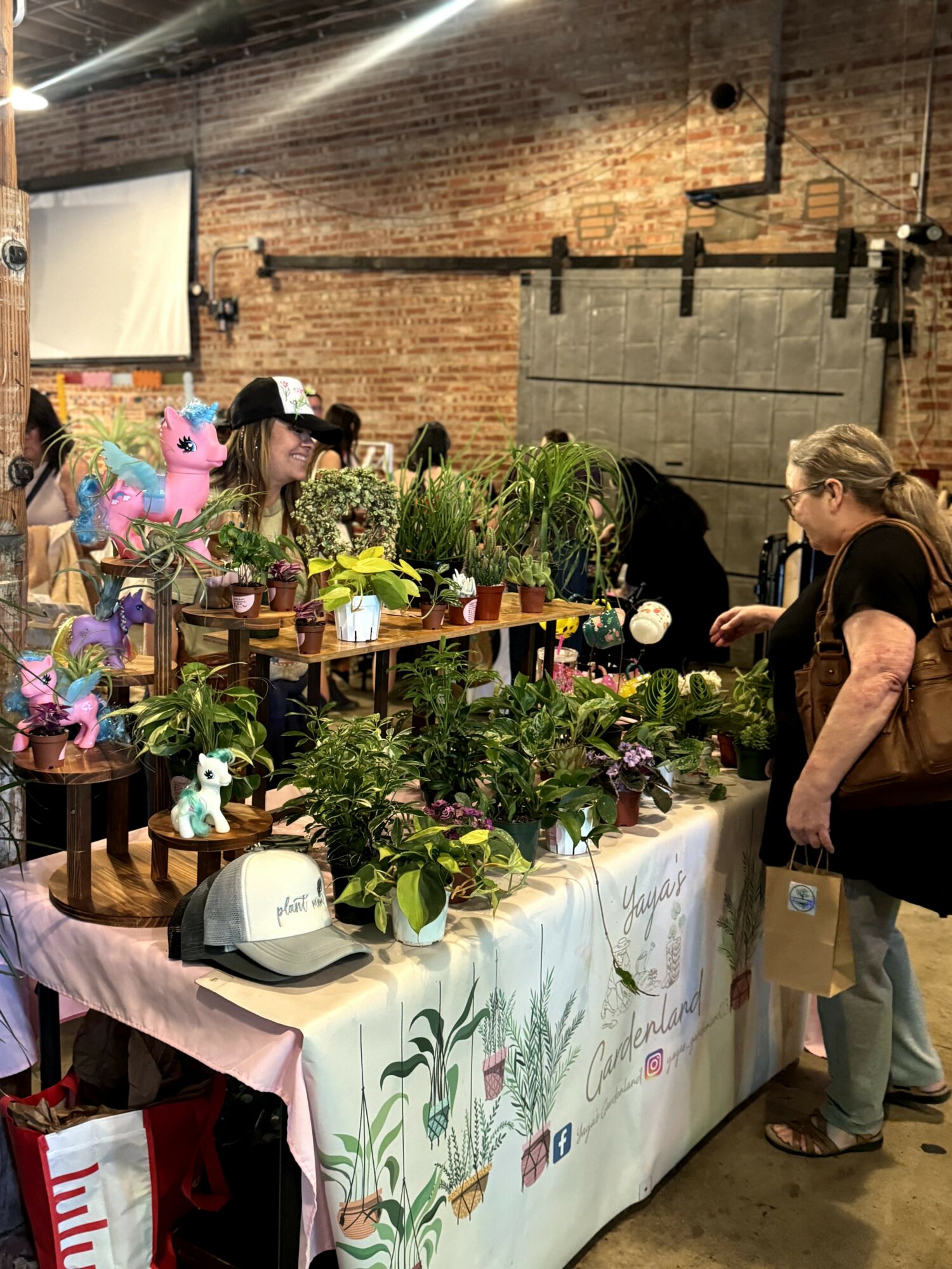 People browsing plants and flowers at an indoor market stall with brick wall background.