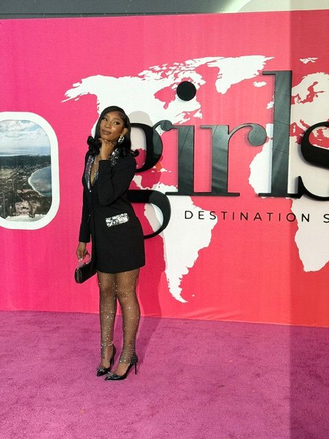 Woman in black dress and blazer standing on pink carpet in front of a pink and white backdrop with text and graphics.