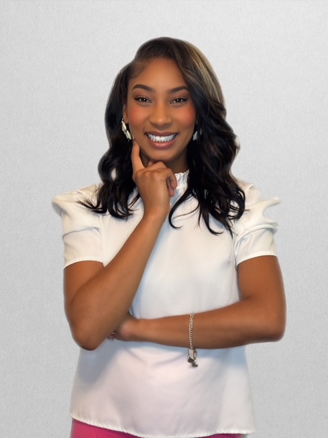 Smiling woman with long dark hair, wearing a white top, posing with her hand near her chin against a plain background.