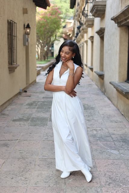 Young woman in white dress standing on sidewalk, holding her hair, with buildings and trees in background.