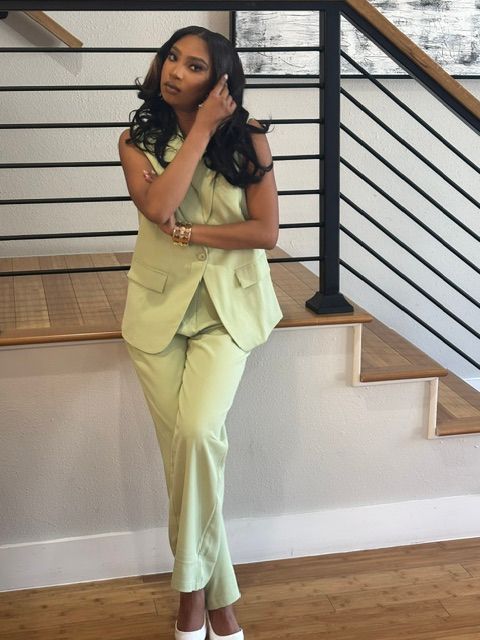 Woman in yellow pants and top poses indoors near staircase, with hand on her face, looking at camera.