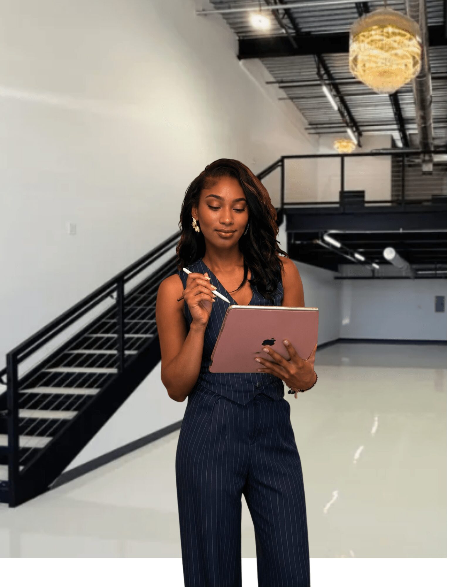 Woman in a dark striped jumpsuit writing in a notebook in a modern indoor space with stairs and ceiling lights.