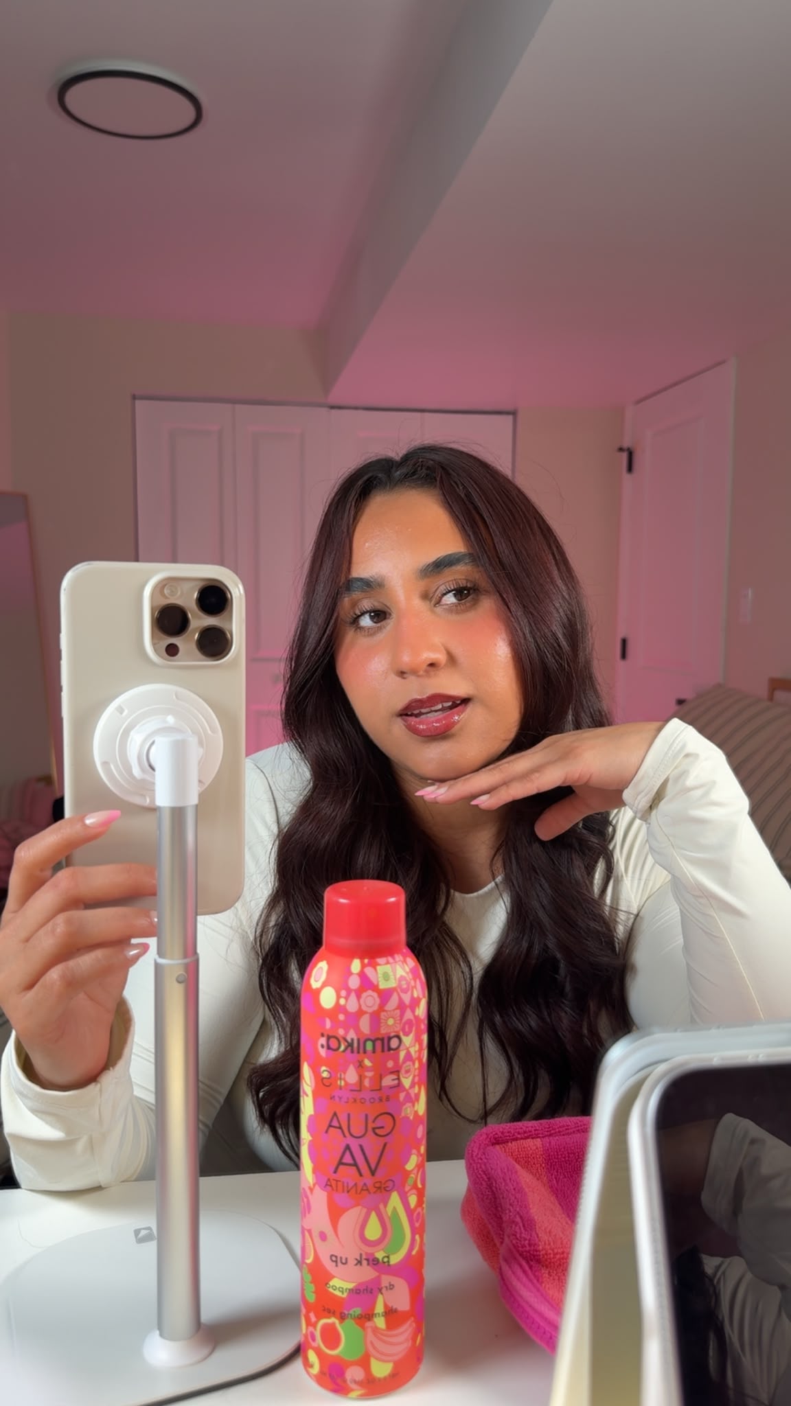 Young woman with long dark hair taking a mirror selfie in a room, holding a phone with a pop socket, with a pink bottle and pink container in front.