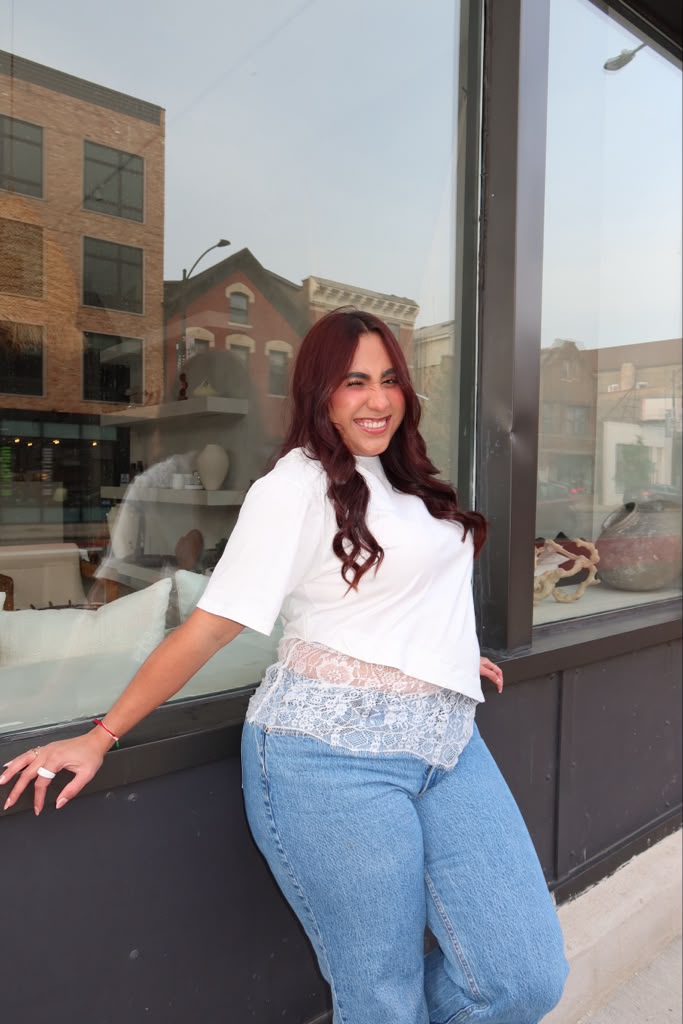 Smiling woman with long wavy hair in white top and blue jeans standing outside near a large window.