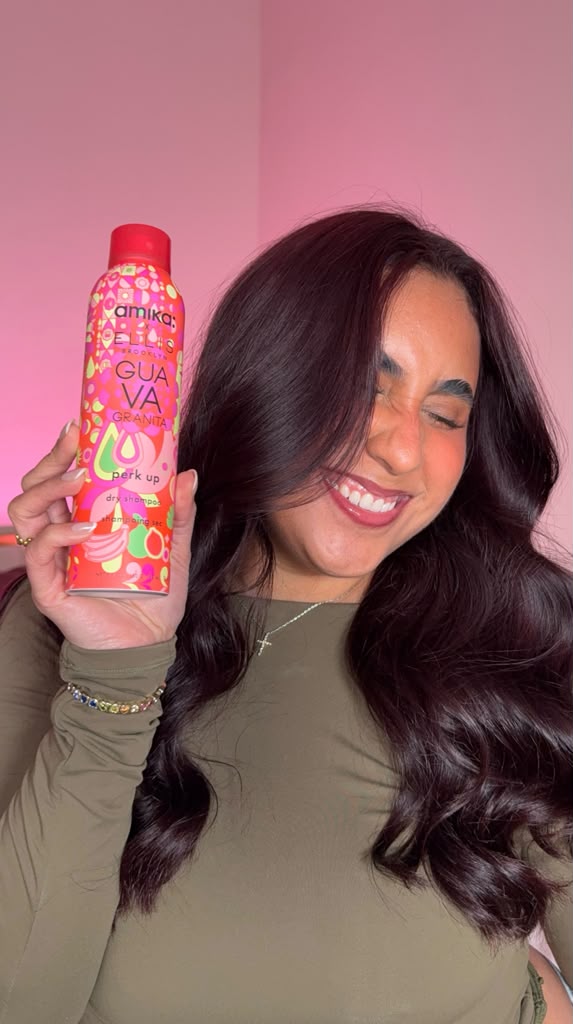 Woman with long dark hair smiling, holding a colorful bottle, against pink background