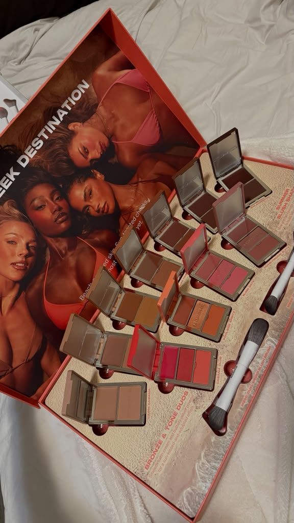 Open makeup palette with multiple shades, a brush, and a promotional card featuring diverse women, on a white surface.