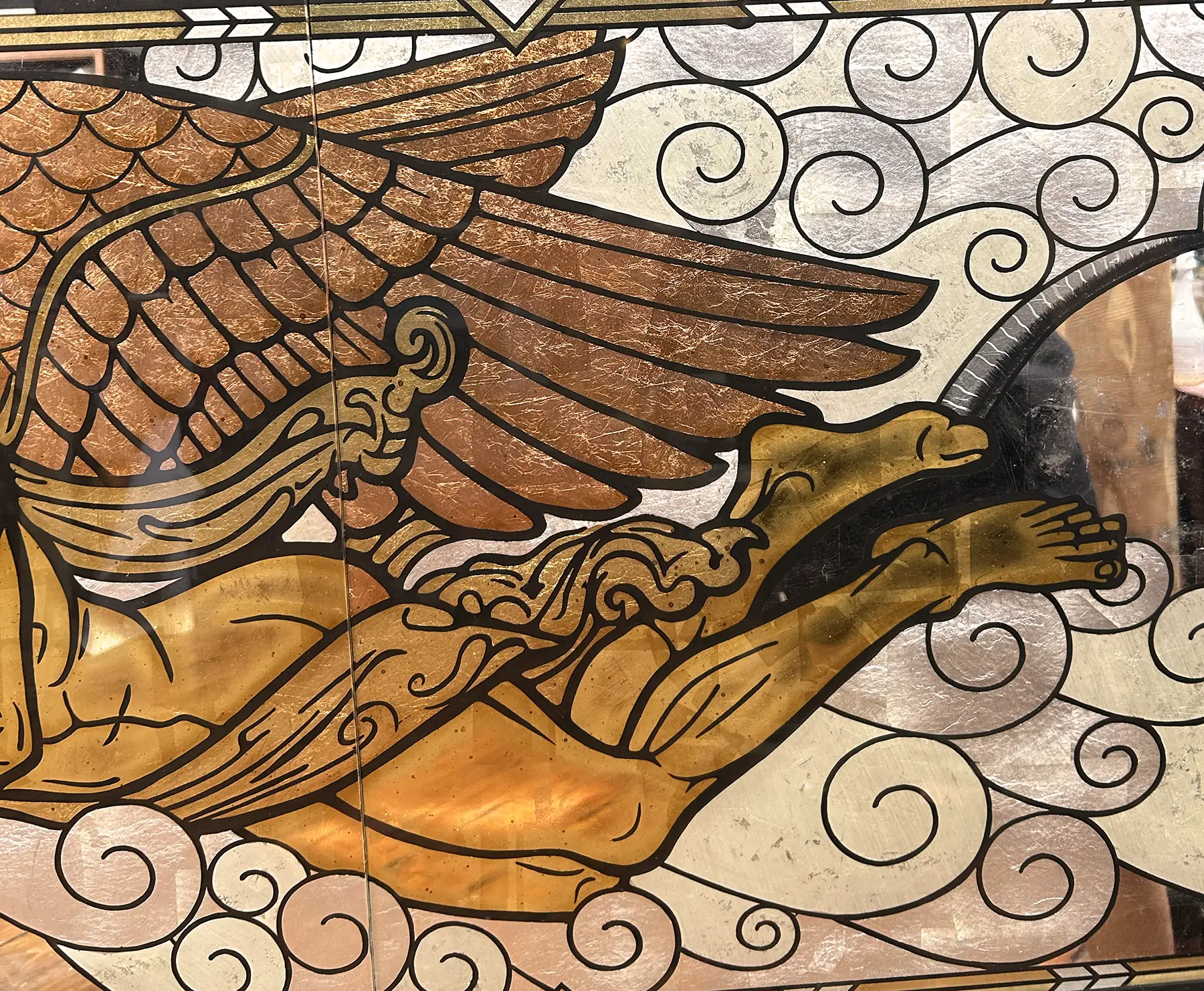 Close-up of a stained glass artwork depicting a winged creature with outstretched wings and a golden body, surrounded by swirling clouds.