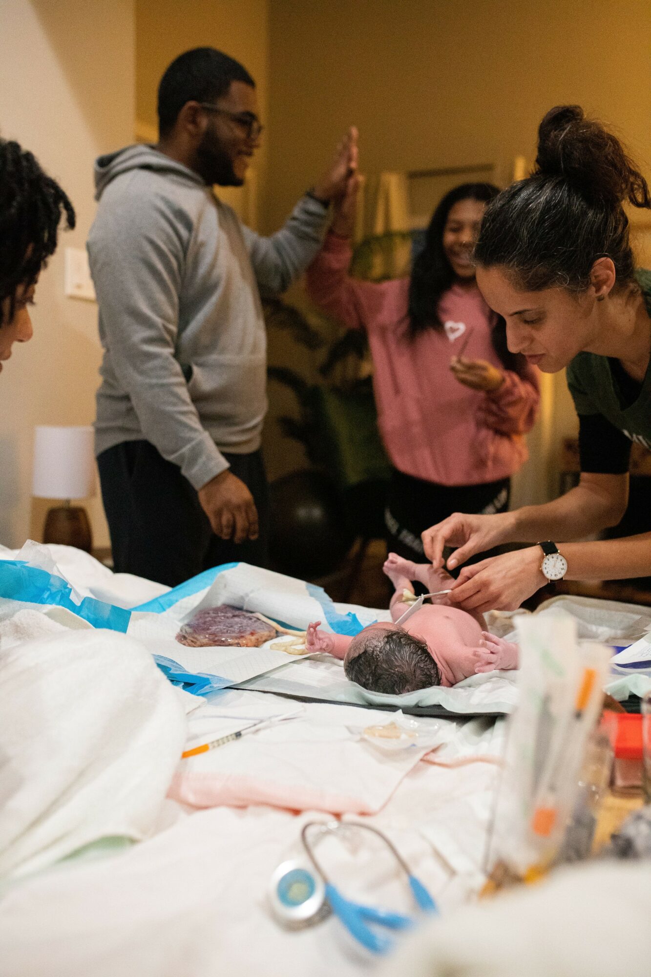 Group of people gathered around a hospital bed with a newborn baby, some giving high-fives, in a hospital room.
