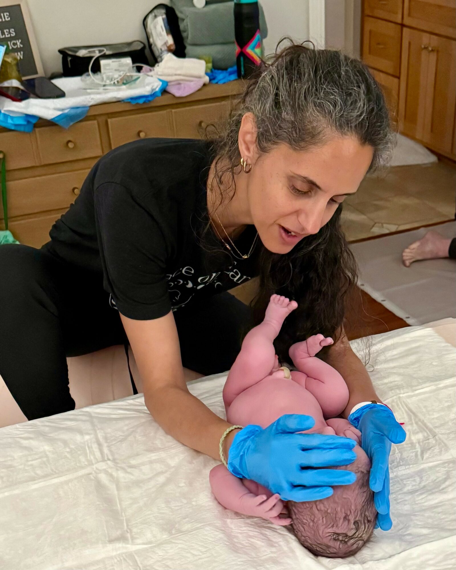 Woman wearing blue gloves holding a newborn baby on a bed, in a room with wooden furniture and cluttered surfaces.