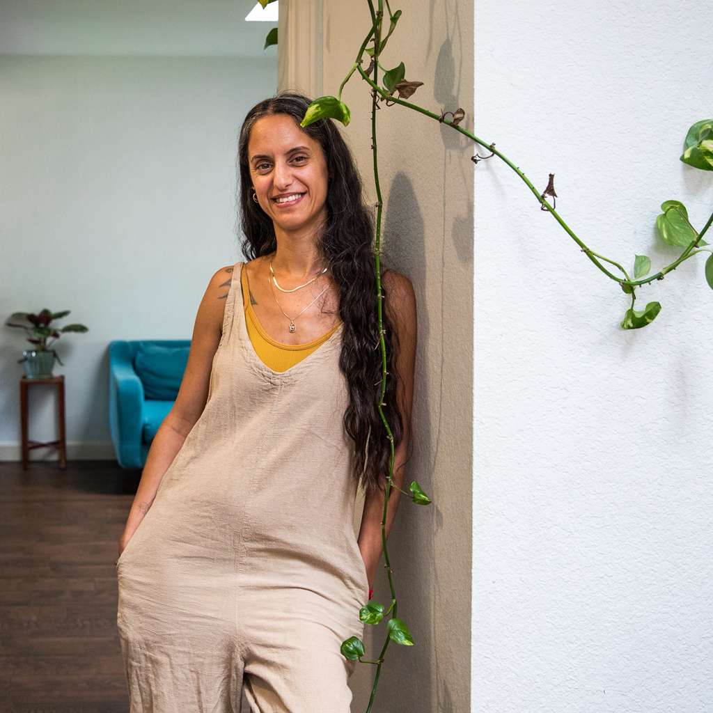 Smiling woman with long dark hair in a beige dress standing indoors near a white wall with a vine plant.