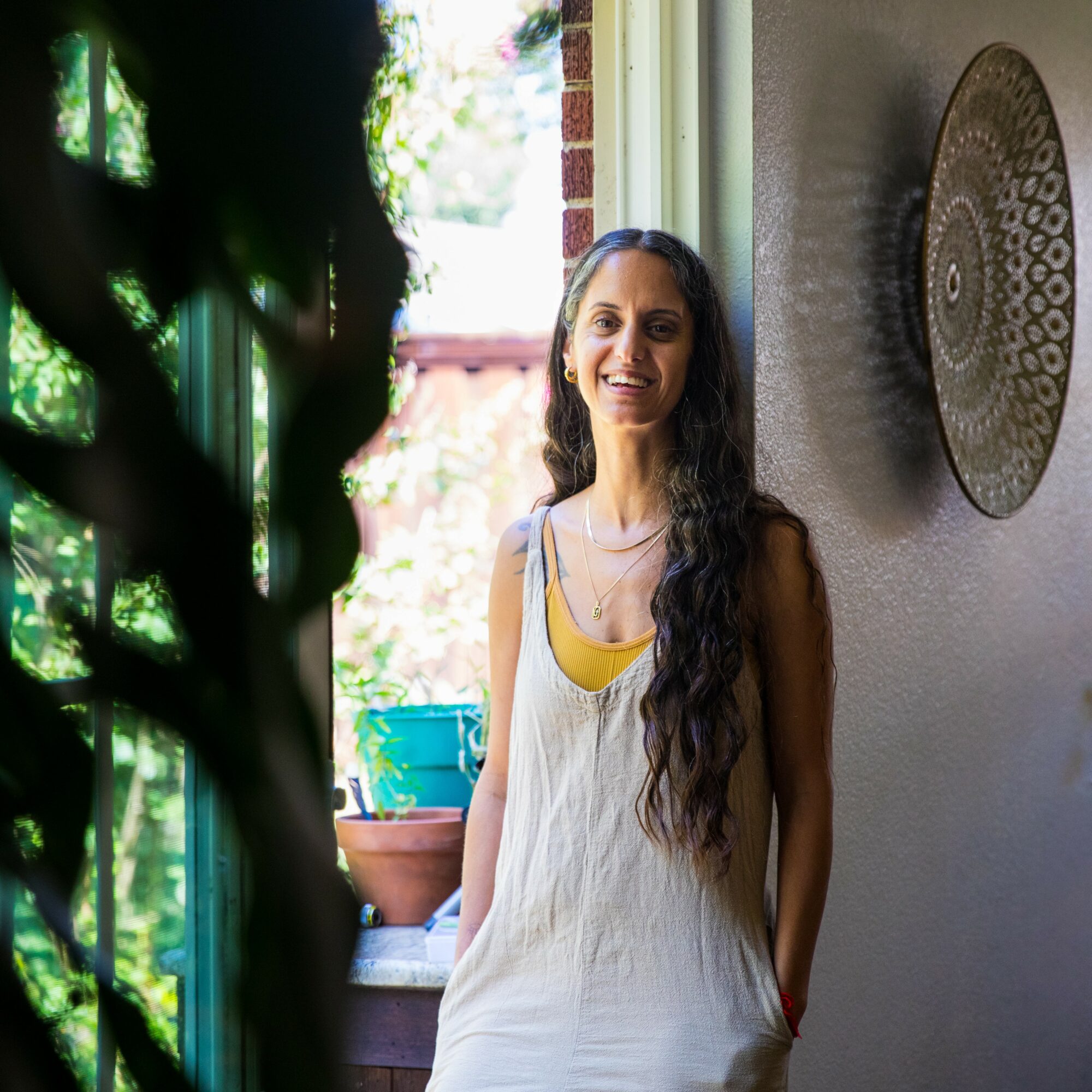 Smiling woman with long dark hair standing indoors near a window with plants outside, decorated wall art behind her.
