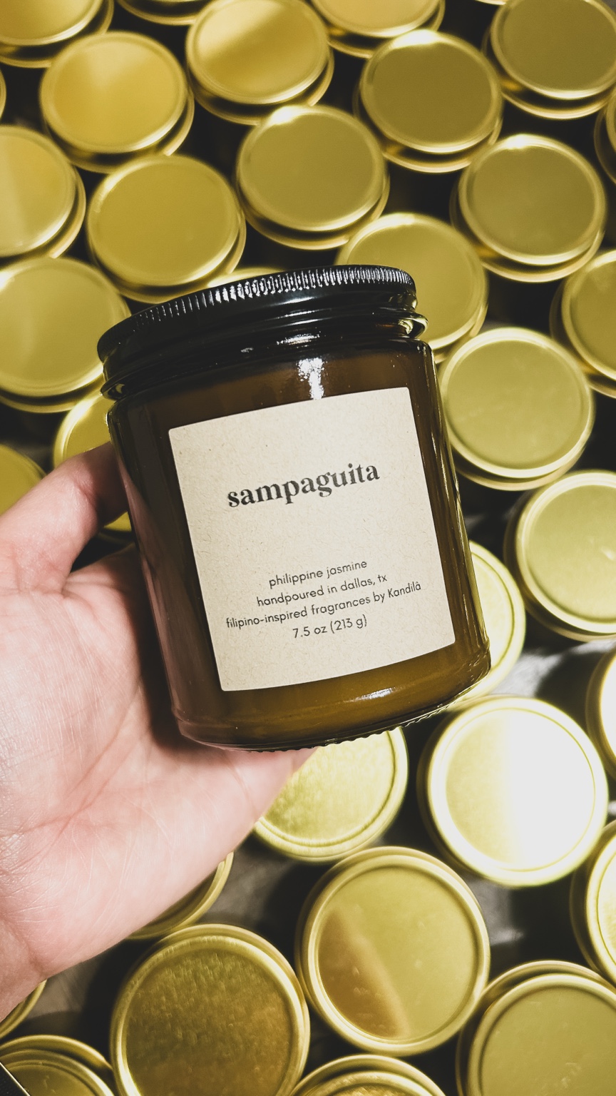 Hand holding a jar labeled 'sampaguira' in front of many gold-colored jars, all with black lids.