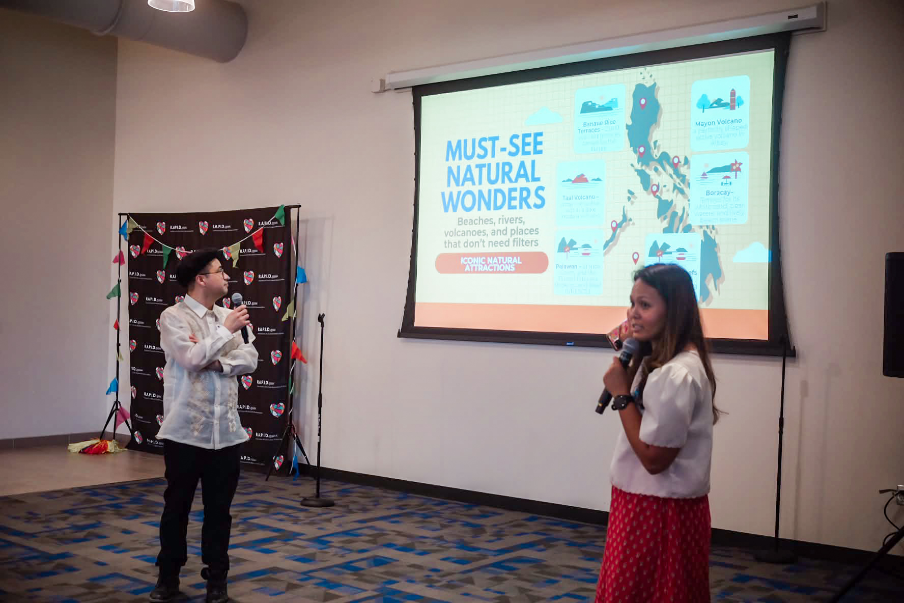 Two women speaking in front of a presentation slide in a room with a patterned carpet and a decorated backdrop.