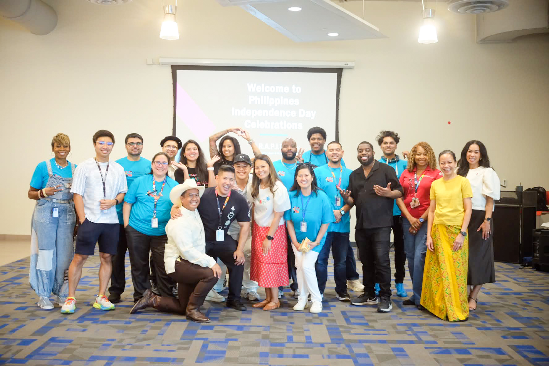 Group of diverse people standing in a conference room, some wearing blue shirts, with a presentation screen behind them.