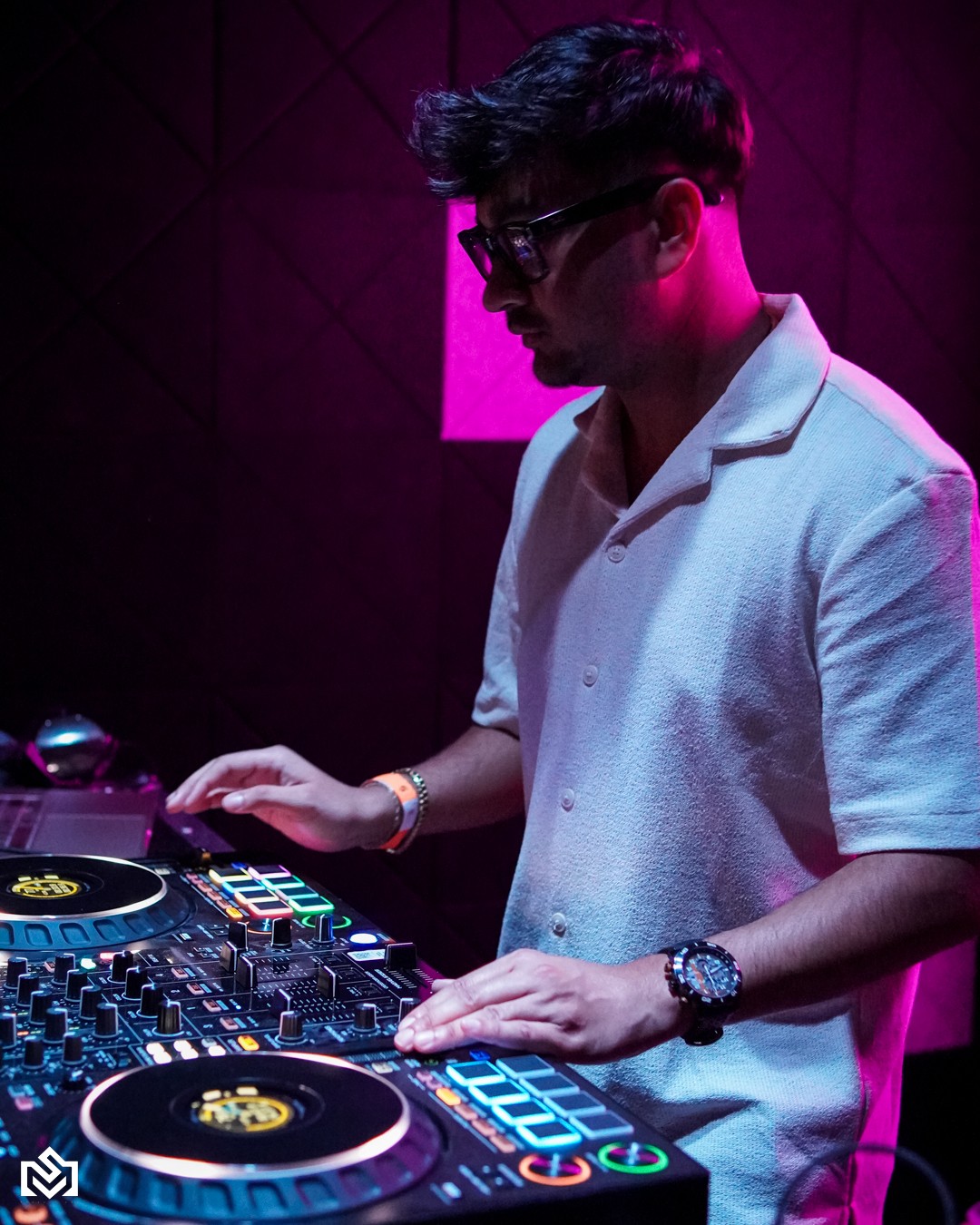 Person with glasses in a white shirt operating DJ equipment in a dimly lit room with pink lighting.
