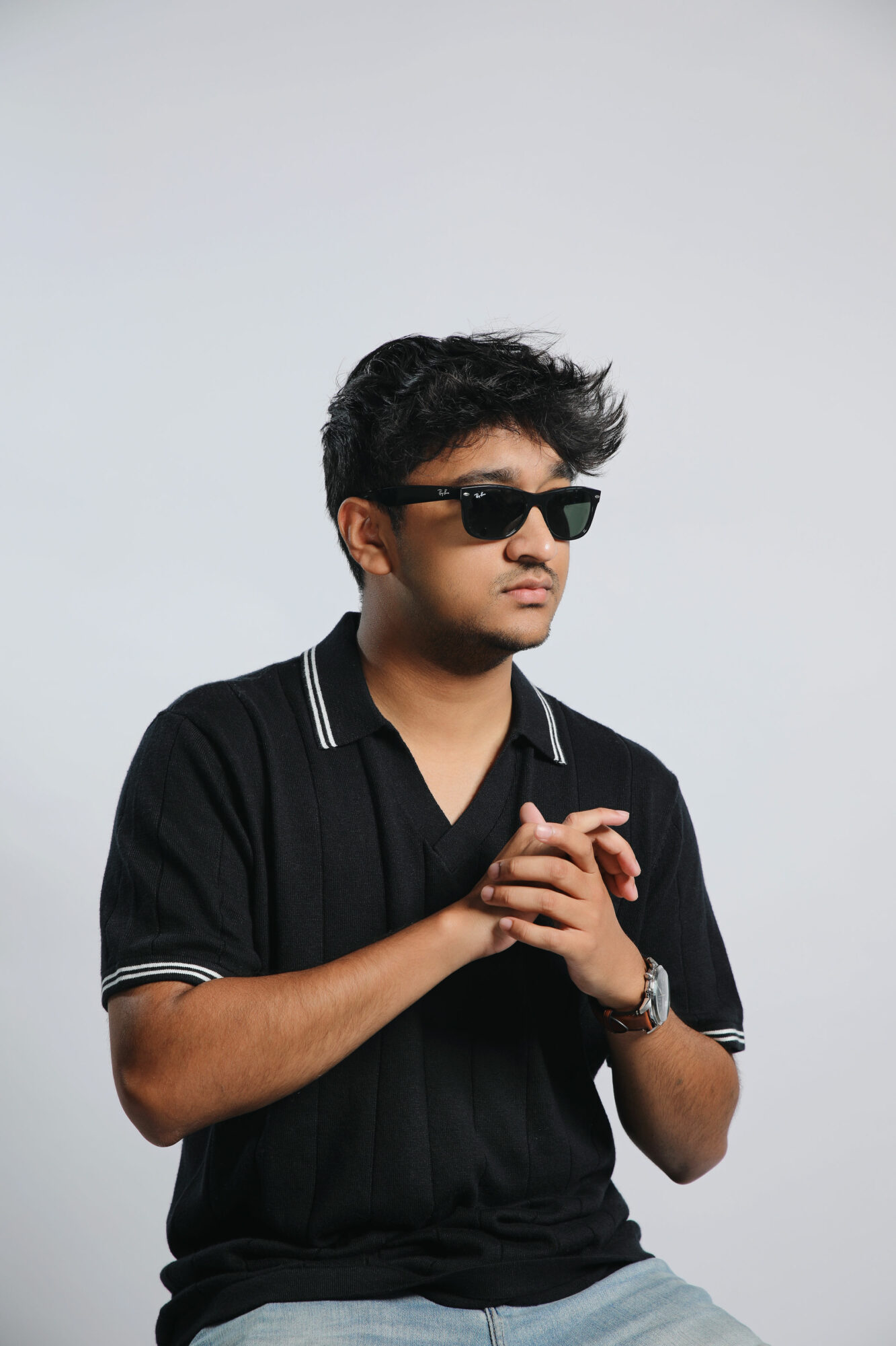 Young man with dark hair wearing sunglasses, black polo shirt, and watch, standing with hands clasped against a plain background.