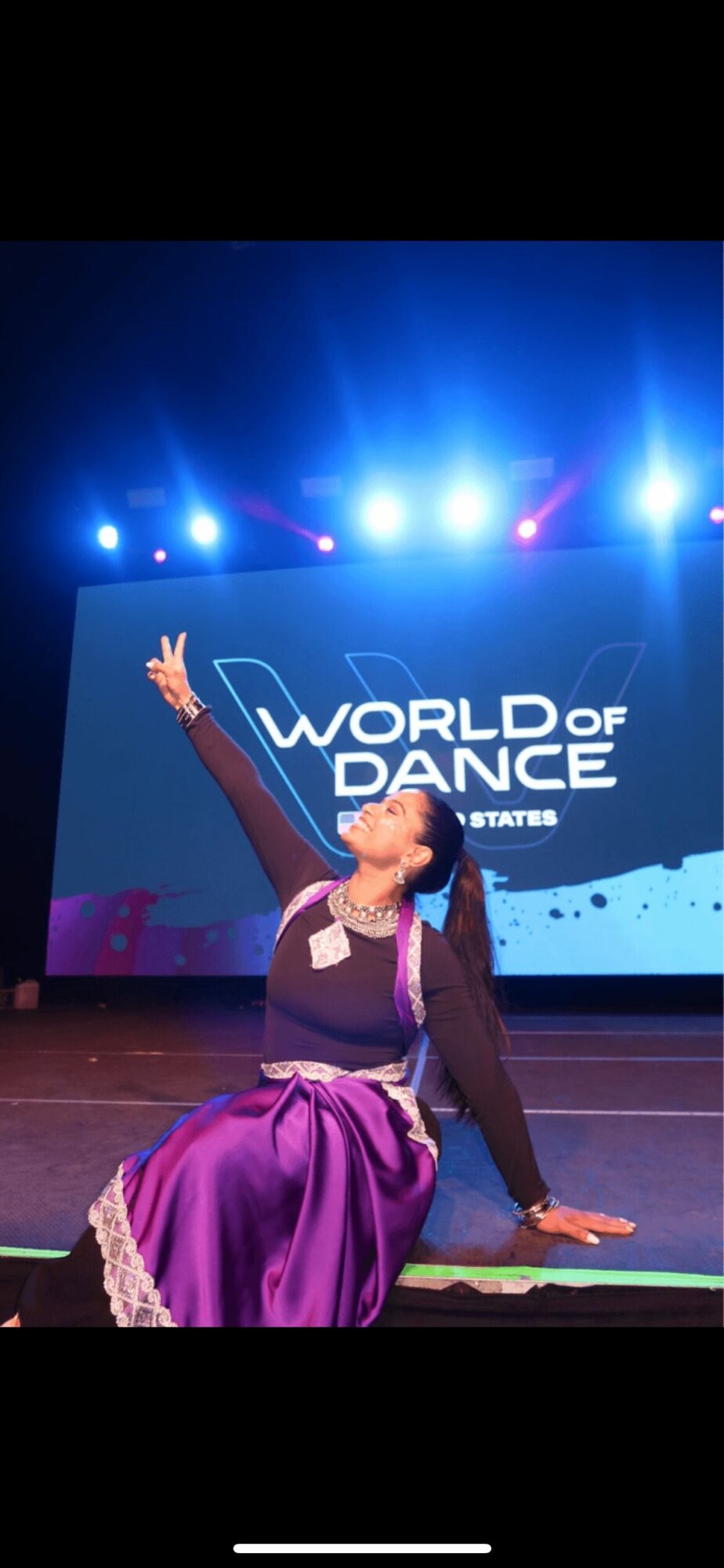 A dancer in a purple costume performs on stage with a 'World of Dance' sign and bright stage lights behind.