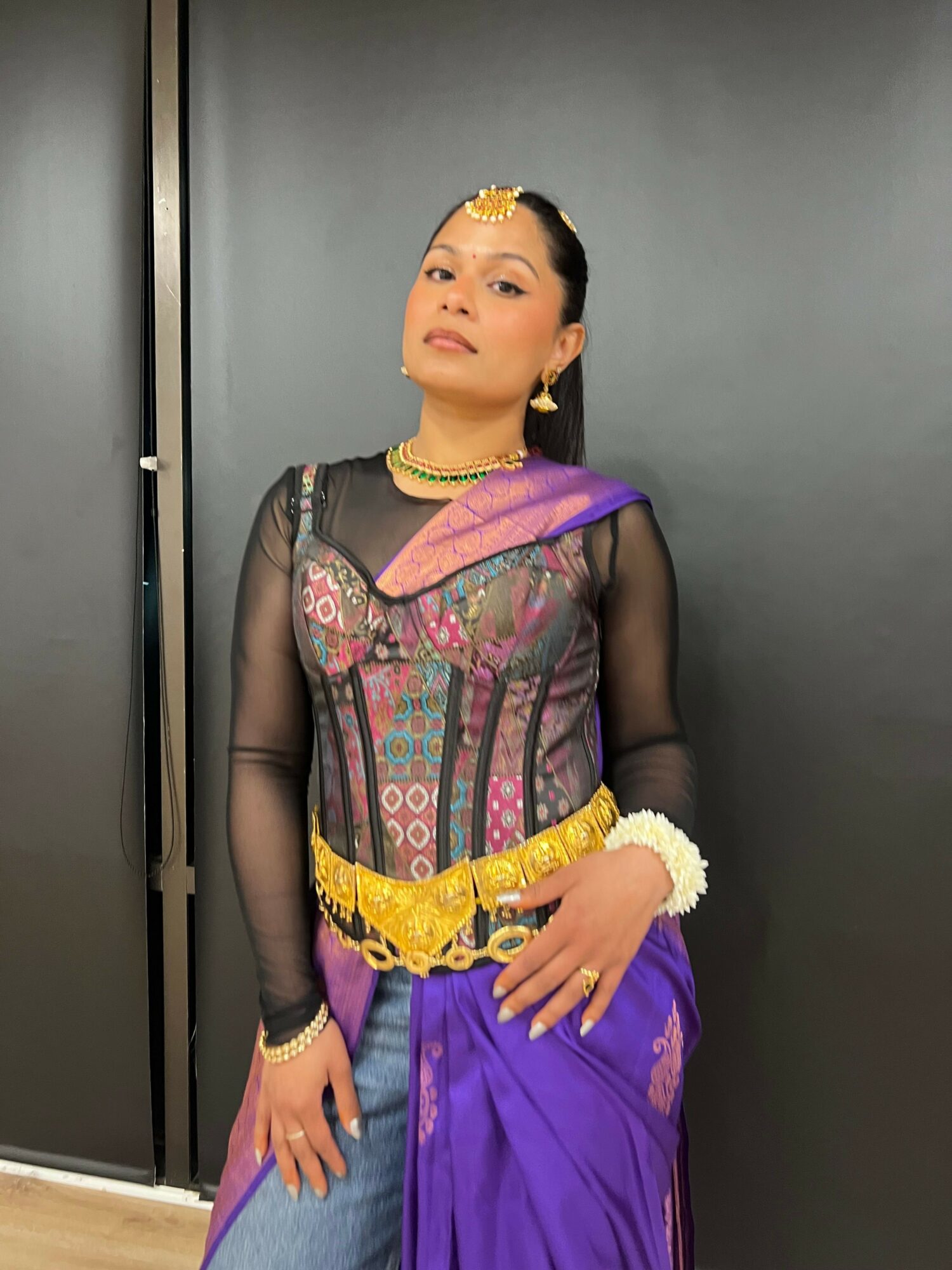 Person in traditional attire with jewelry, standing against a gray background, looking at the camera.