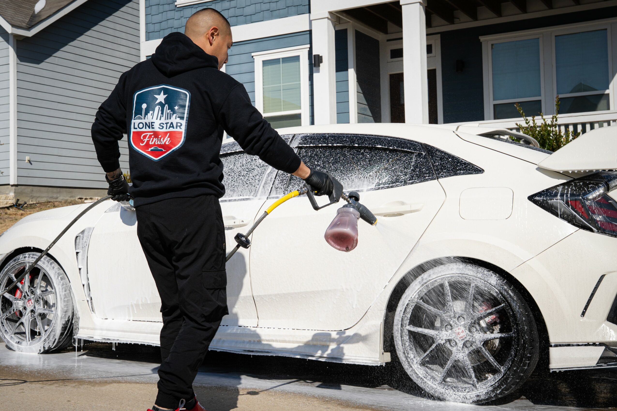 Person cleaning a white car with a foam brush, wearing a black hoodie with a logo on the back, in front of a house.