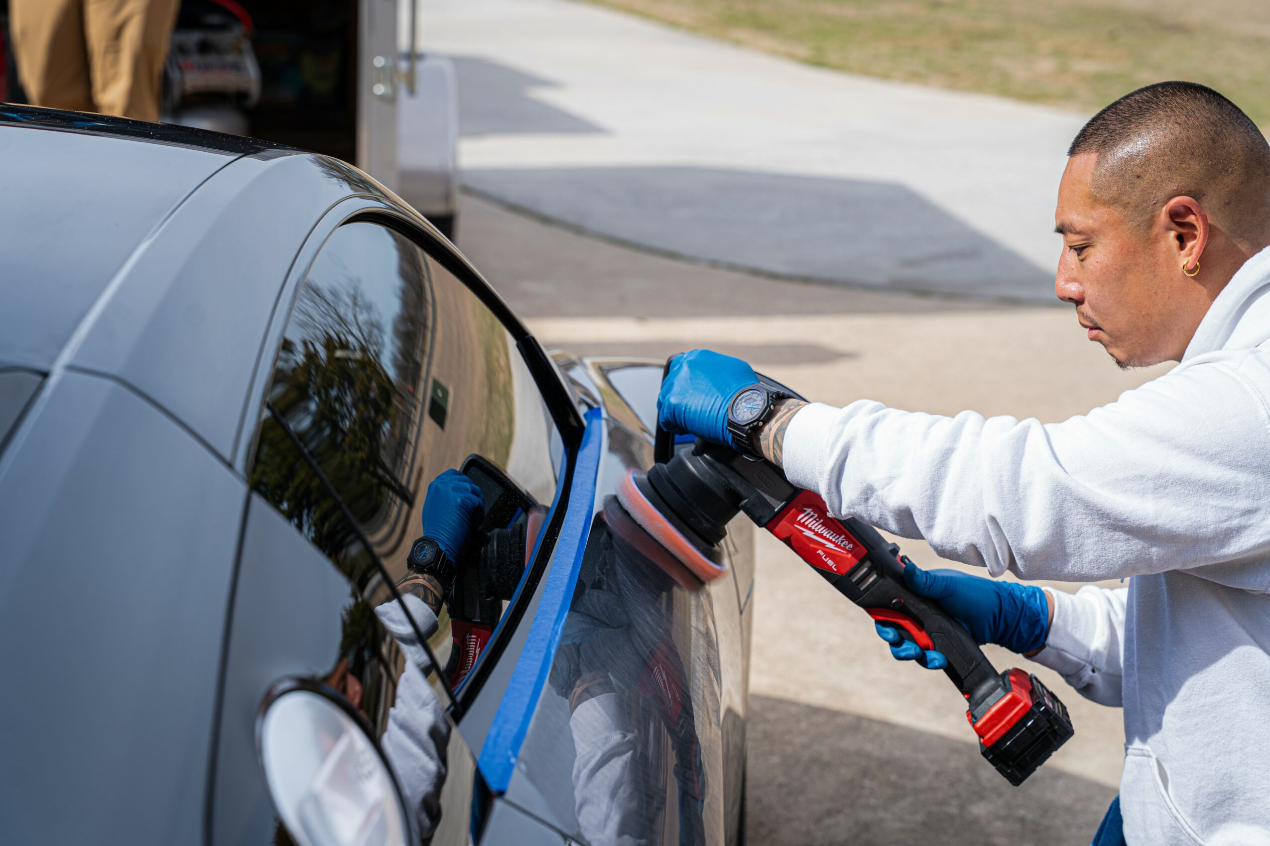 Person cleaning car window with a spray and cloth, wearing gloves, outdoors on a sunny day.