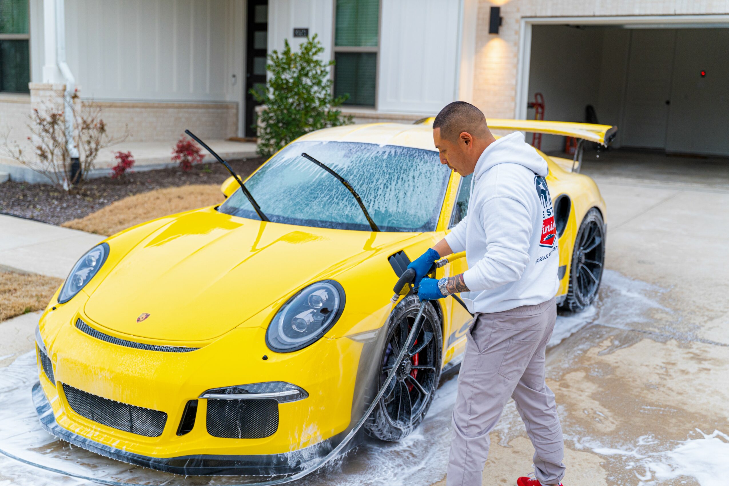 Person washing a yellow sports car with a hose in driveway, house in background.