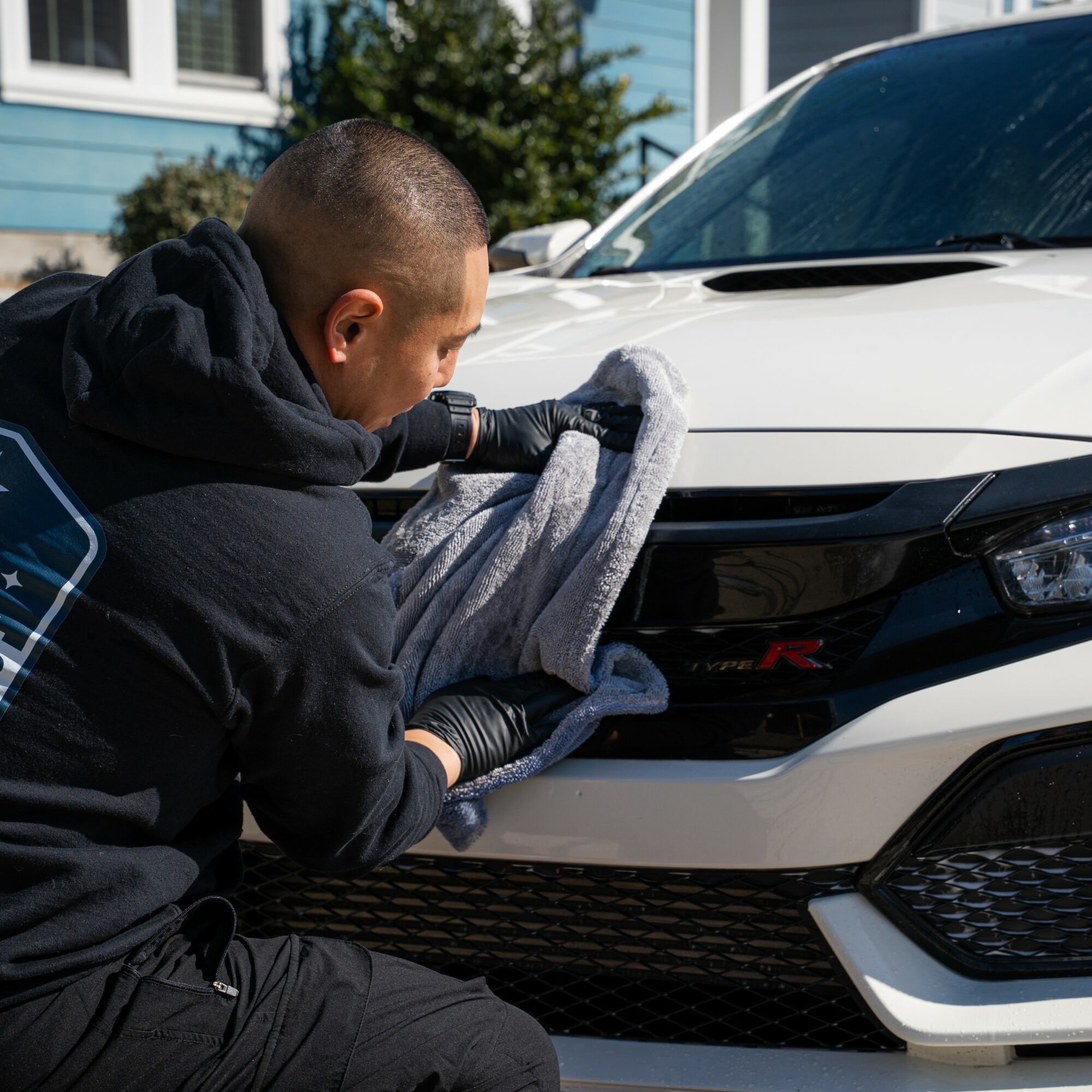 Person cleaning front of white car with a cloth, outdoor setting, house and trees in background.