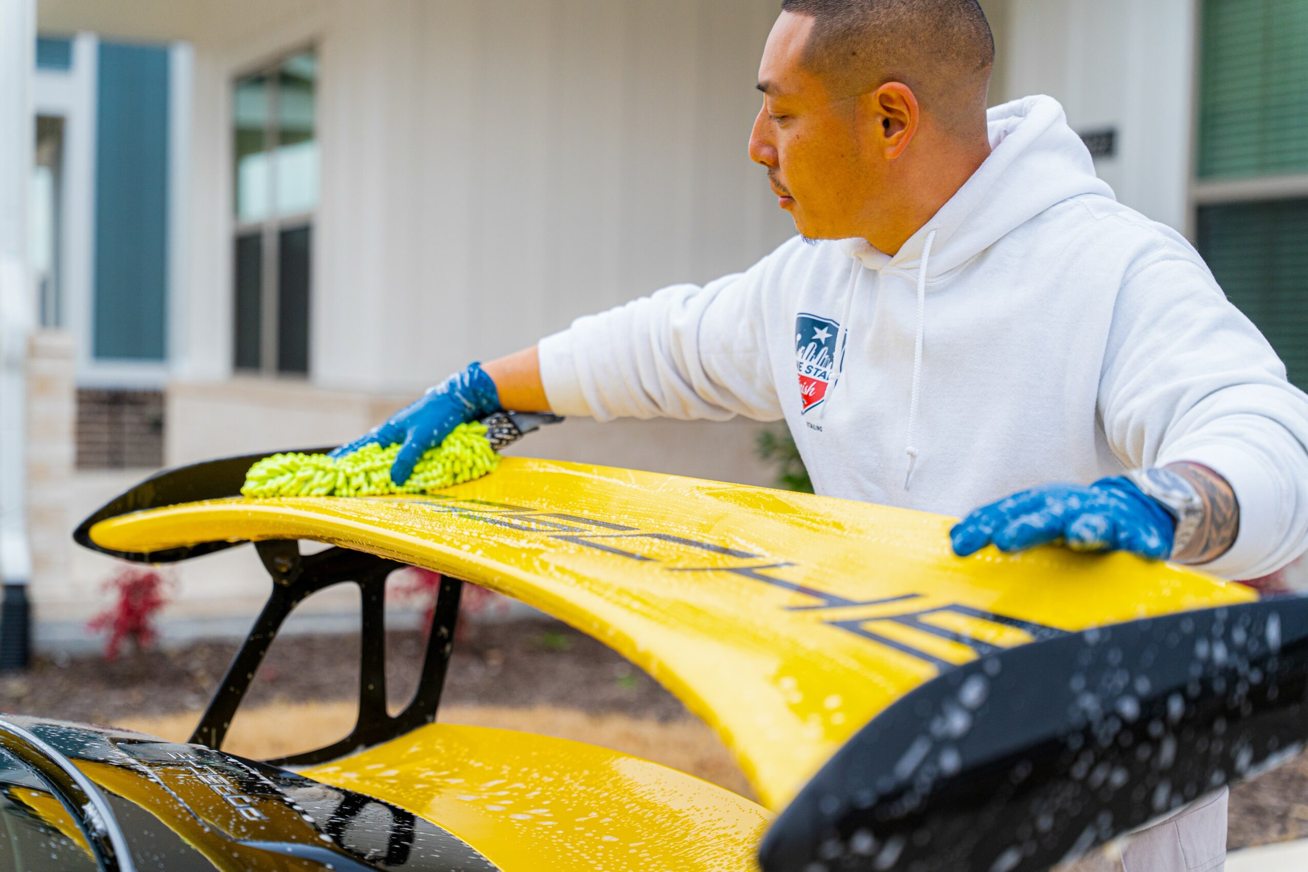 Person cleaning a yellow surfboard outdoors, wearing a white hoodie and blue gloves.