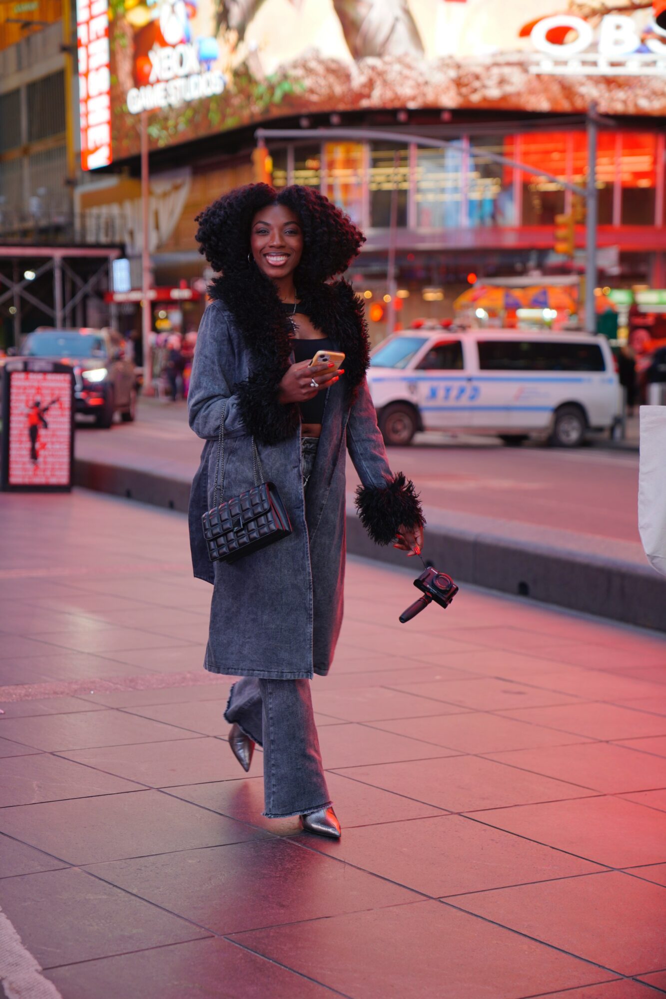 Woman with curly hair in a long coat holding a camera and phone, smiling on city street at dusk.