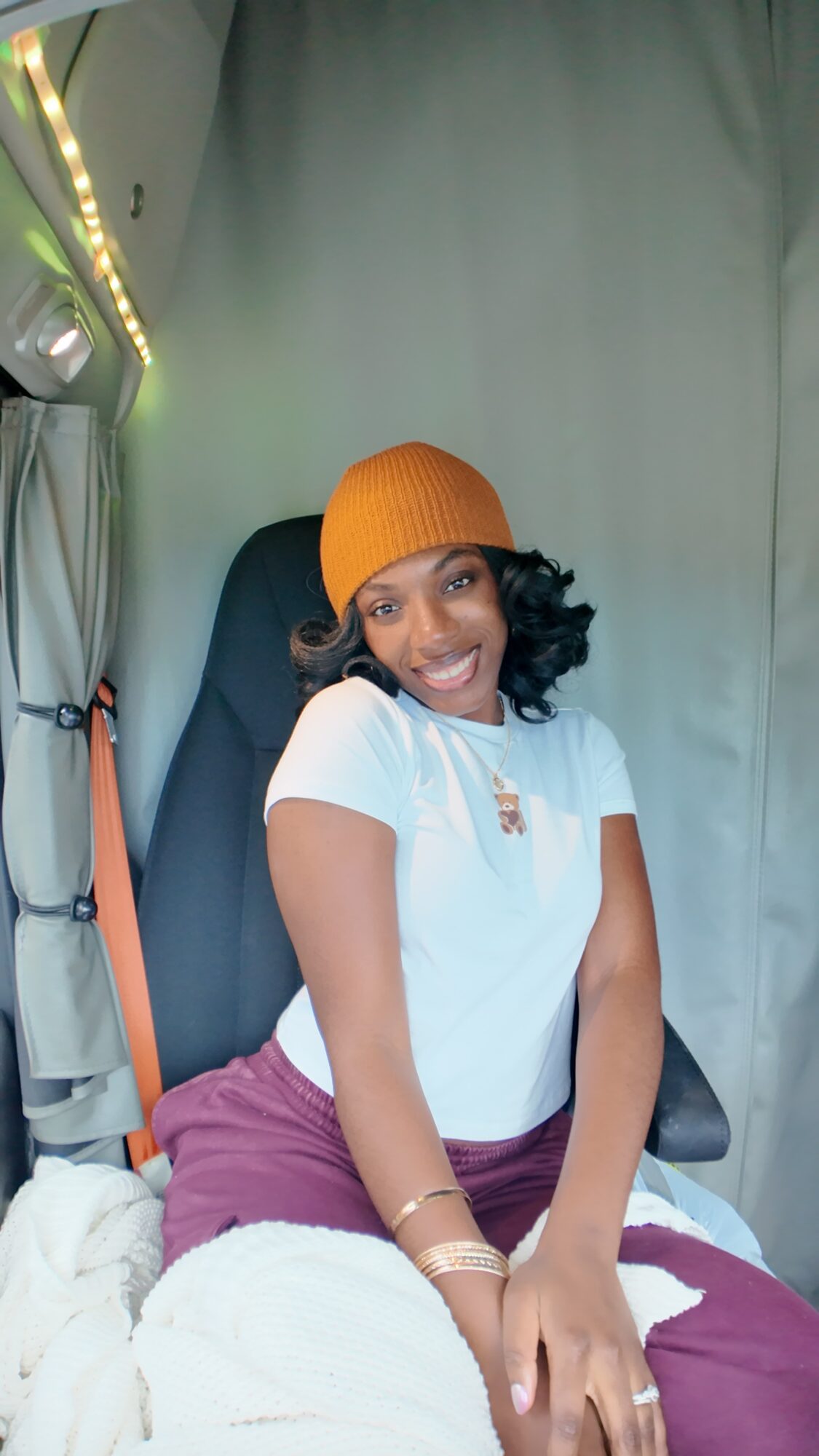 Smiling woman wearing an orange beanie and white top, sitting on a bed with a gray curtain background.